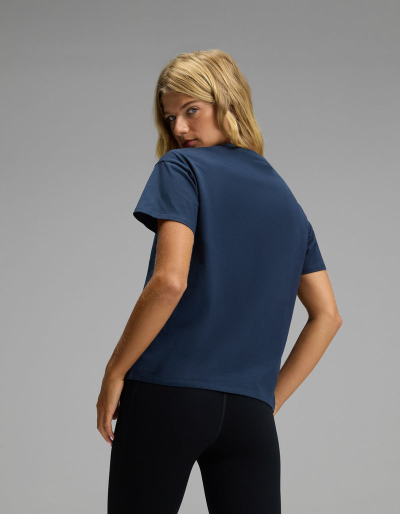 A woman with blonde hair stands with her back to the camera, turning her head to look over her shoulder while wearing Cozy Earth’s Women’s All Day Tee in navy blue and black pants, set against a plain gray background.