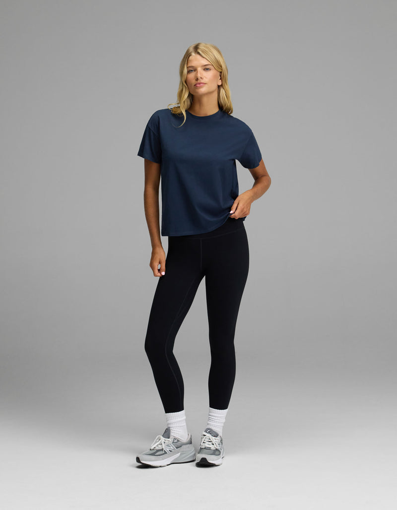 A person wearing the Cozy Earth Women’s All Day Tee in navy, black leggings, white socks, and gray sneakers stands against a plain gray background with one hand on their hip.