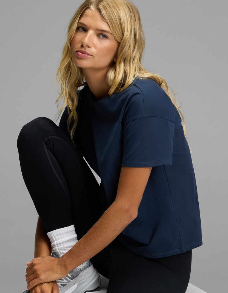 A person with long blonde hair wears the Cozy Earth Women’s All Day Tee in navy, paired with black leggings and white sneakers, sitting with one knee up against a plain gray background.