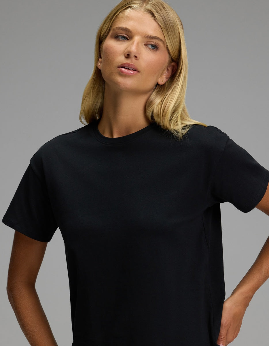 A woman with long blonde hair wears the Women’s All Day Tee by Cozy Earth in black and gazes slightly to the side against a plain gray background.|Color:Jet Black