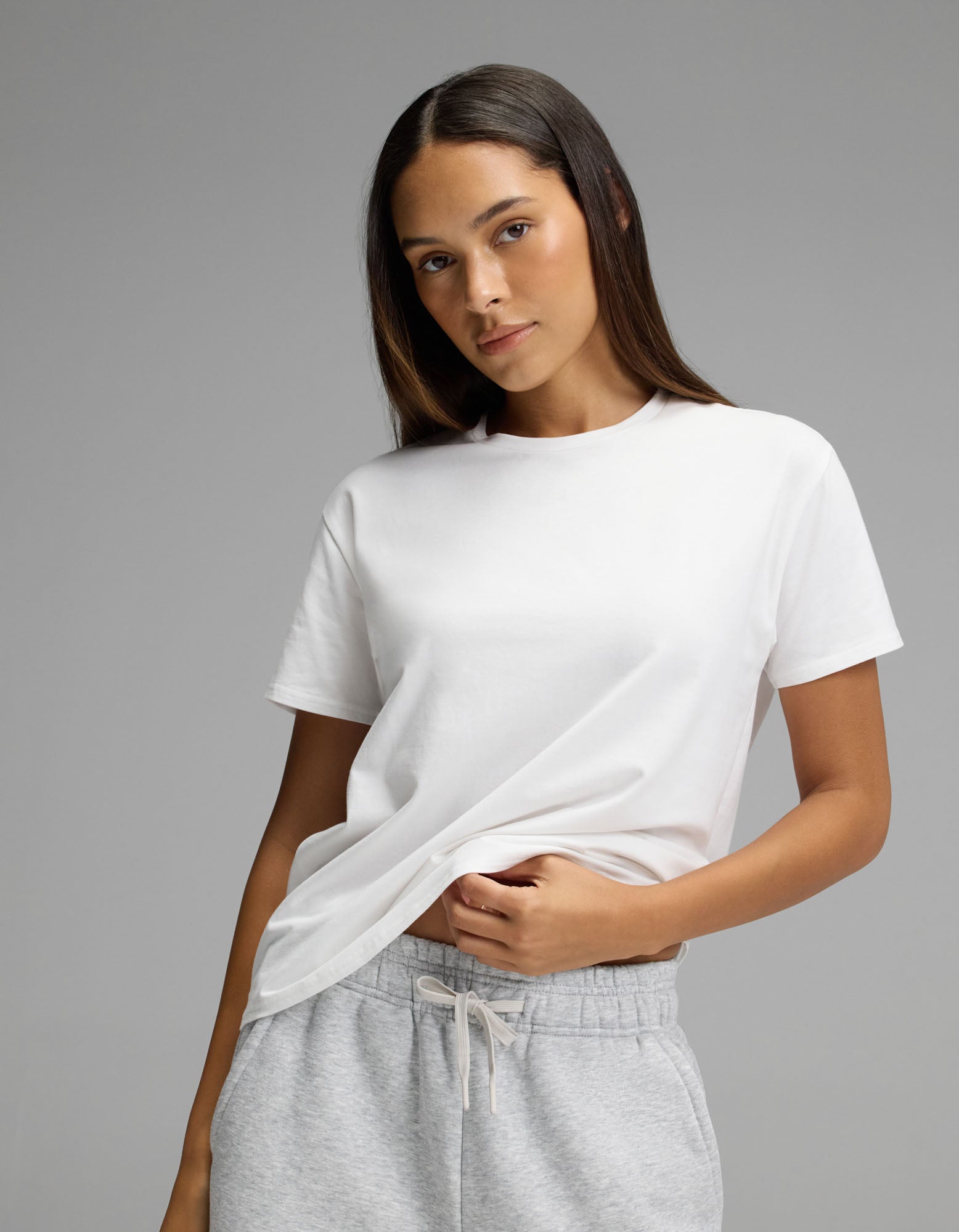 A woman wears the Cozy Earth Women’s All Day Tee in plain white and light gray sweatpants, standing against a neutral background. She looks at the camera with a slight smile while holding the hem of her shirt.