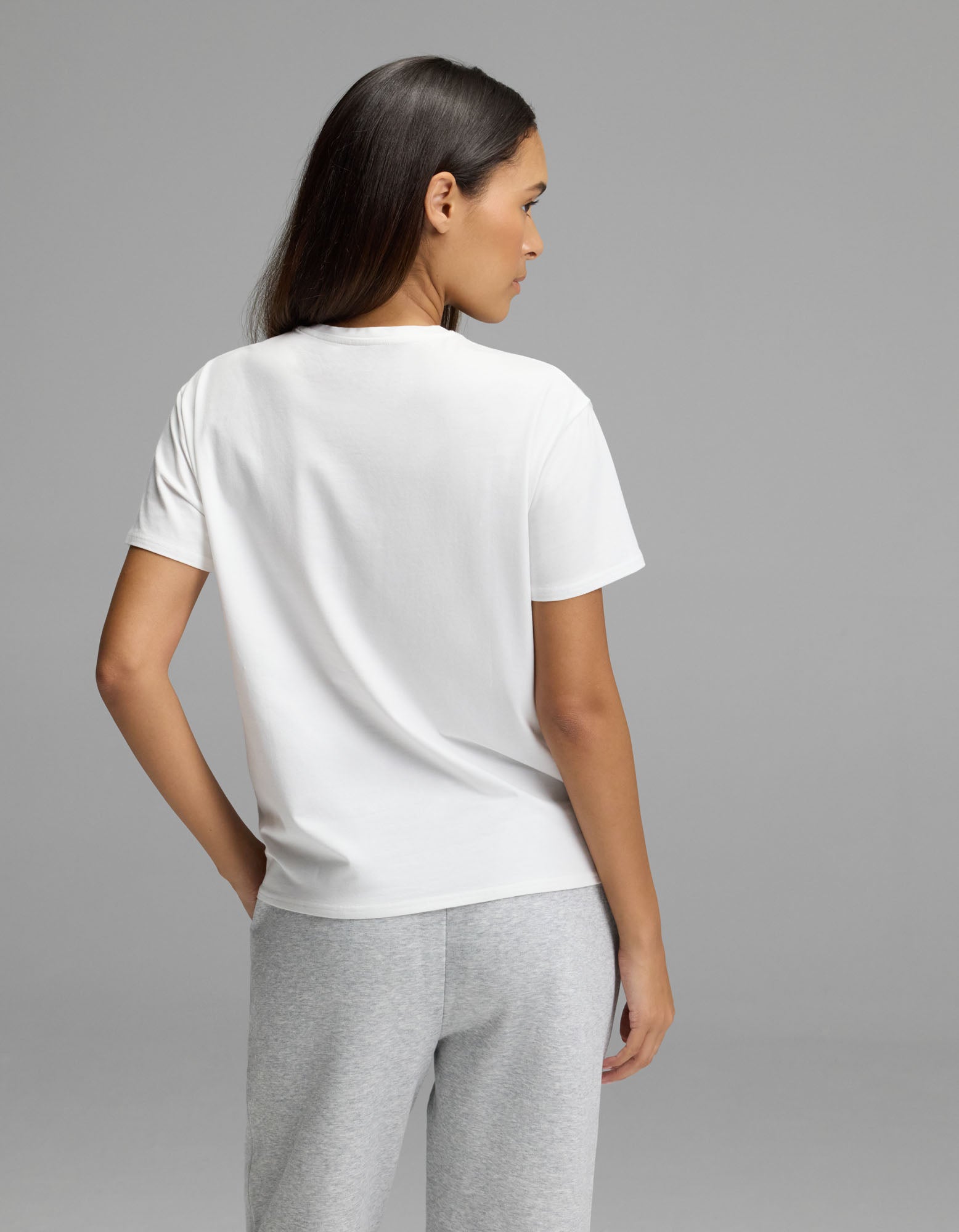 A woman with long dark hair wears the Cozy Earth Women’s All Day Tee in white and gray pants, standing against a plain gray background and looking to the side.|Color:White