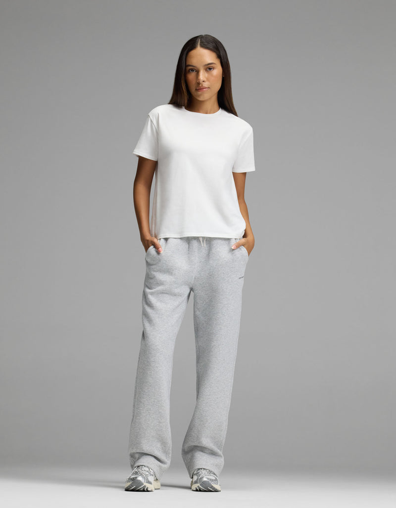 A woman stands facing forward against a plain gray background, wearing the Cozy Earth Women’s All Day Tee in white, light gray sweatpants, and sneakers, with her hands in her pockets.