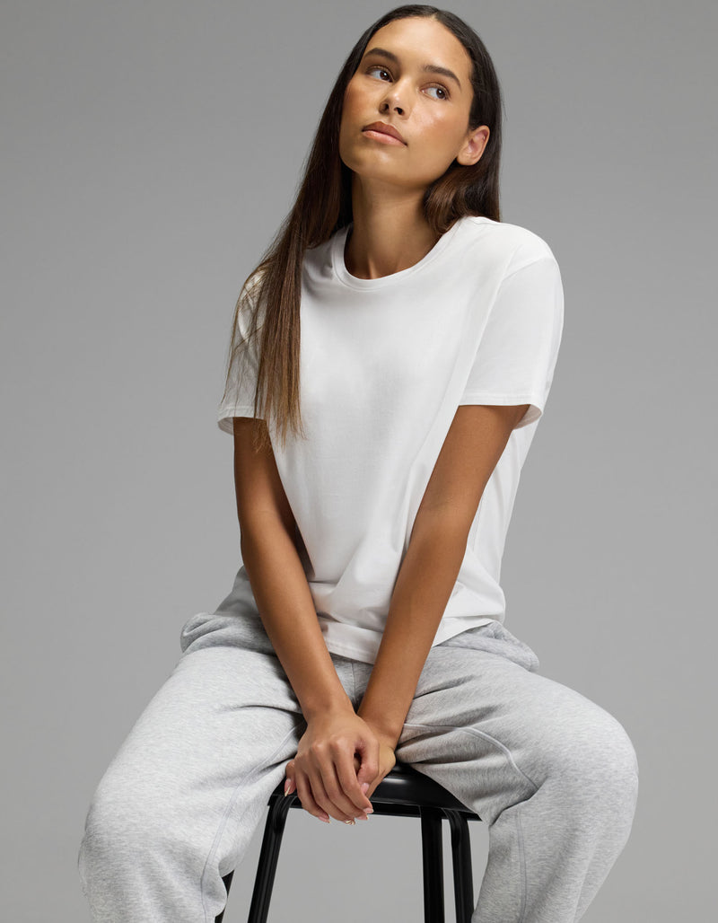 A woman wearing the Cozy Earth Women’s All Day Tee in white and gray sweatpants sits on a black stool against a plain gray background, looking slightly up and to the side with a relaxed expression.