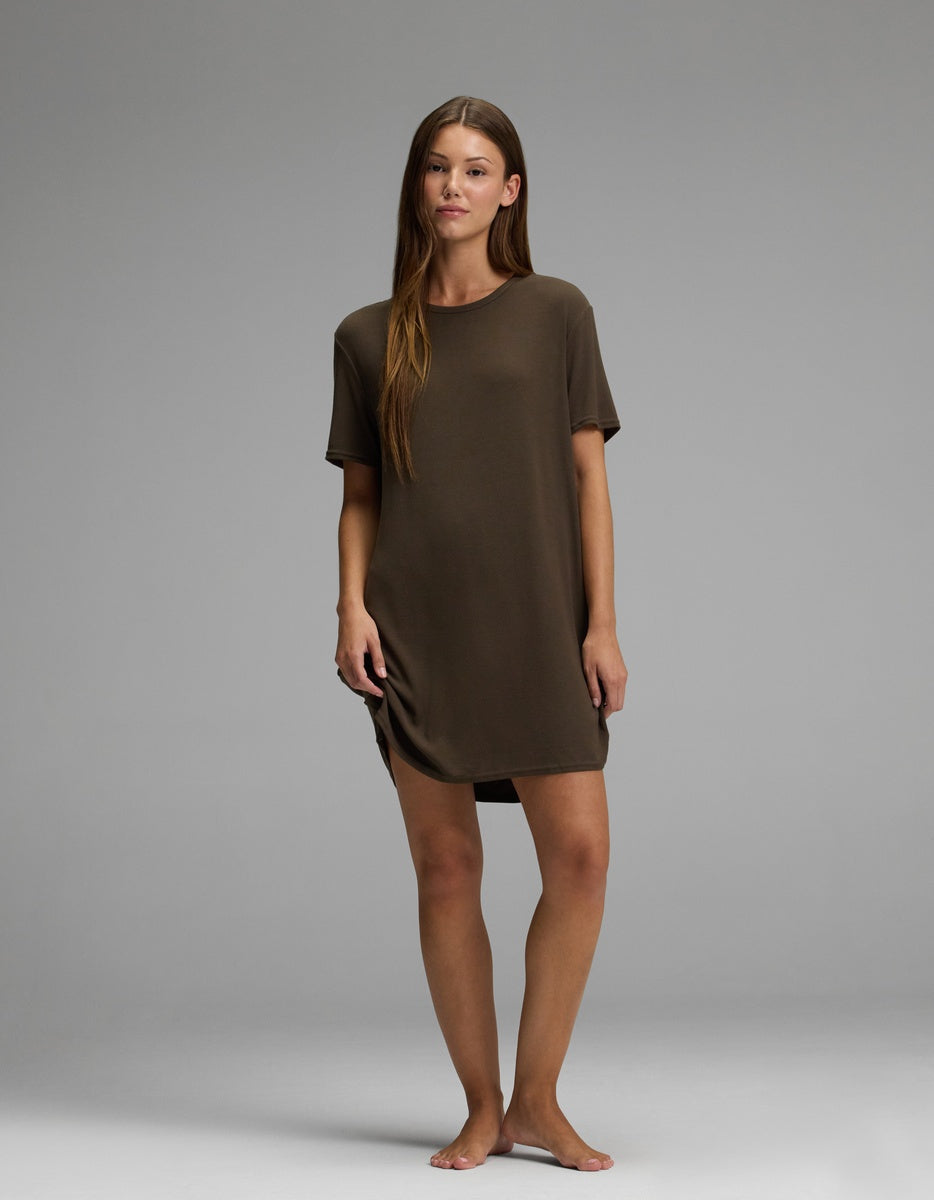 A woman stands barefoot on a plain backdrop, wearing the Cozy Earth Women’s Bamboo Rib-Knit Boyfriend Sleep Dress in brown. She faces forward with a relaxed posture and a neutral expression.
