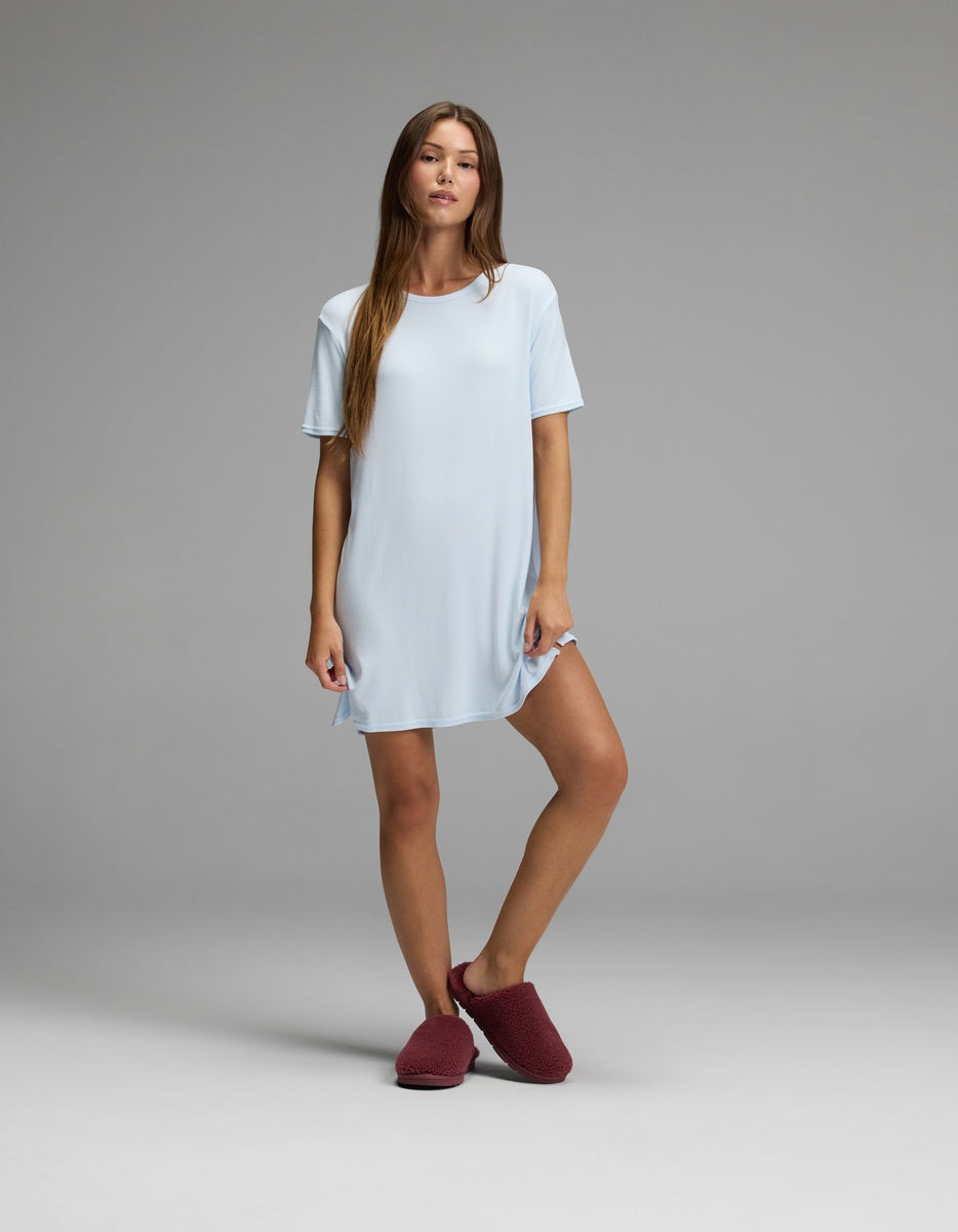 A woman stands against a gray background, wearing the Cozy Earth Women’s Bamboo Rib-Knit Boyfriend Sleep Dress in light blue paired with maroon slippers. She has long brown hair, a relaxed neutral expression, and her left knee is slightly bent.