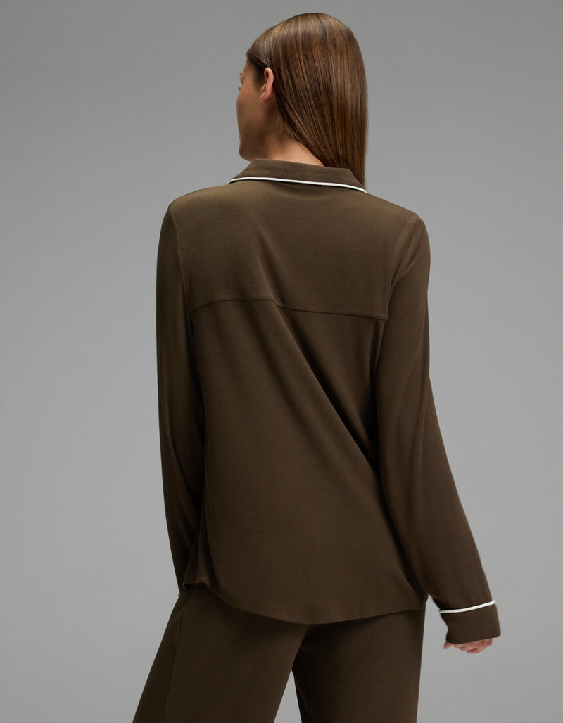 A person with straight brown hair is seen from behind wearing a Cozy Earth Women’s Bamboo Rib-Knit Classic Long Sleeve Pajama Top in brown, featuring white piping along the collar and cuffs, paired with matching pants. The background is plain and gray.