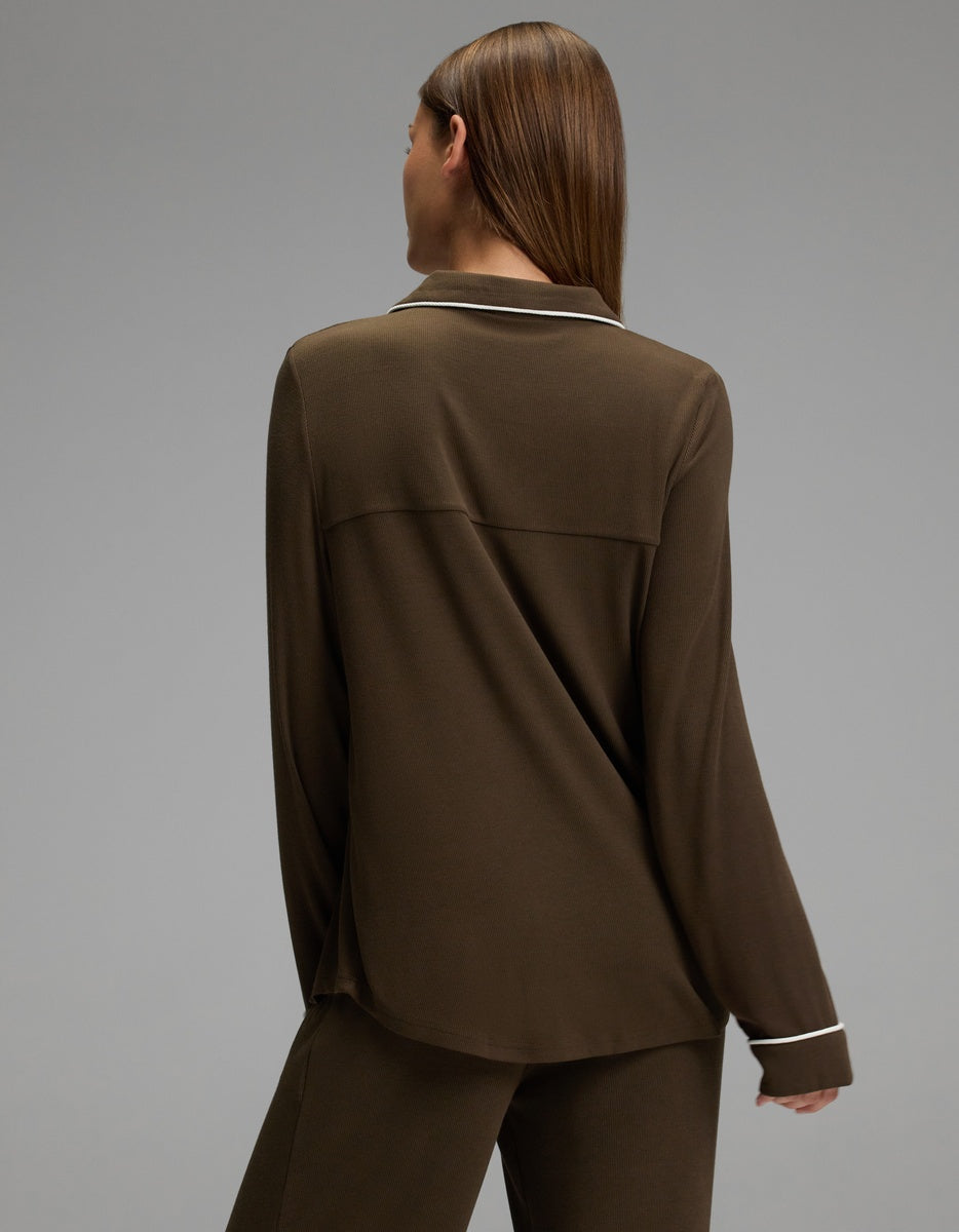 A person with straight brown hair is seen from behind wearing a Cozy Earth Women’s Bamboo Rib-Knit Classic Long Sleeve Pajama Top in brown, featuring white piping along the collar and cuffs, paired with matching pants. The background is plain and gray.|Color:Espresso