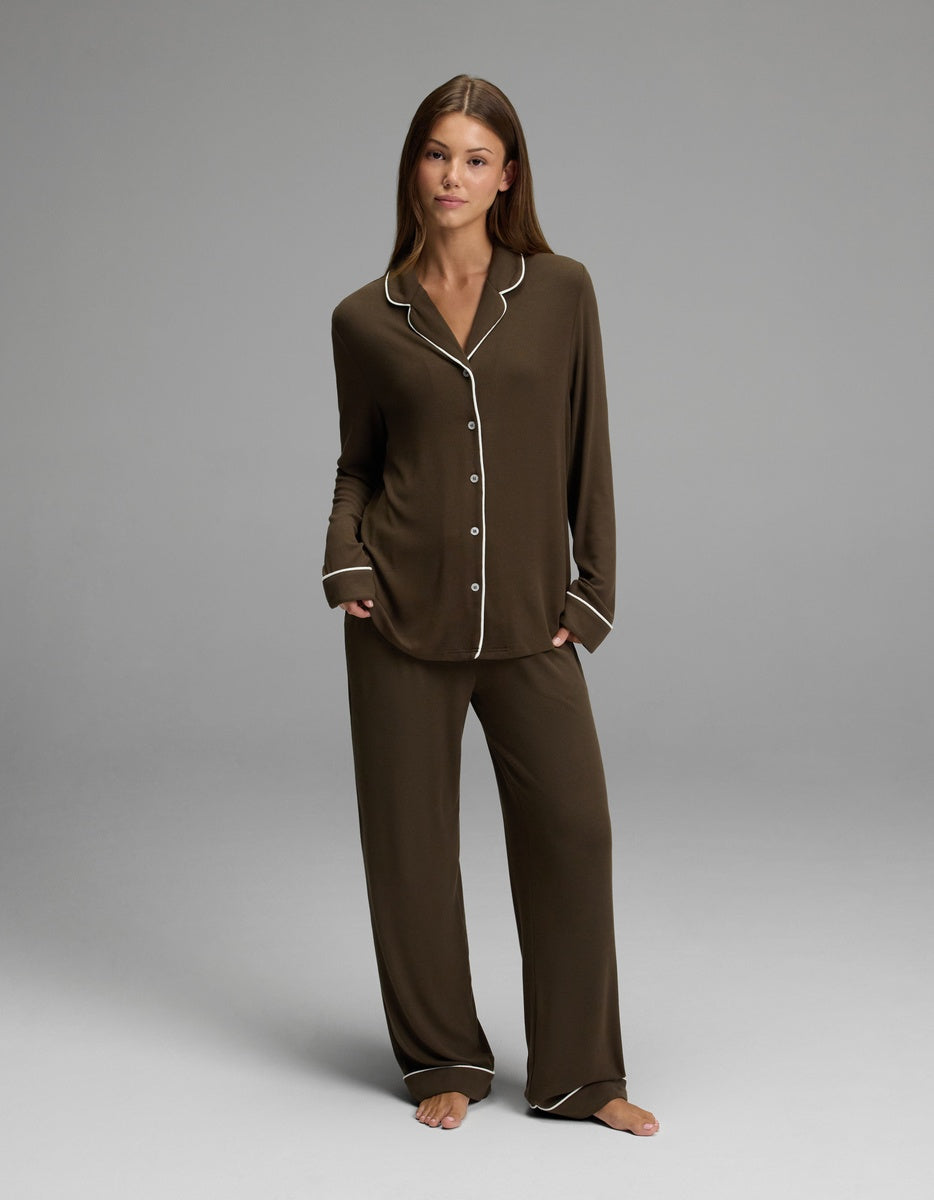 A woman stands barefoot on a gray background, looking at the camera with a neutral expression while wearing the Cozy Earth Women’s Bamboo Rib-Knit Classic Long Sleeve Pajama Top in dark brown with white trim.