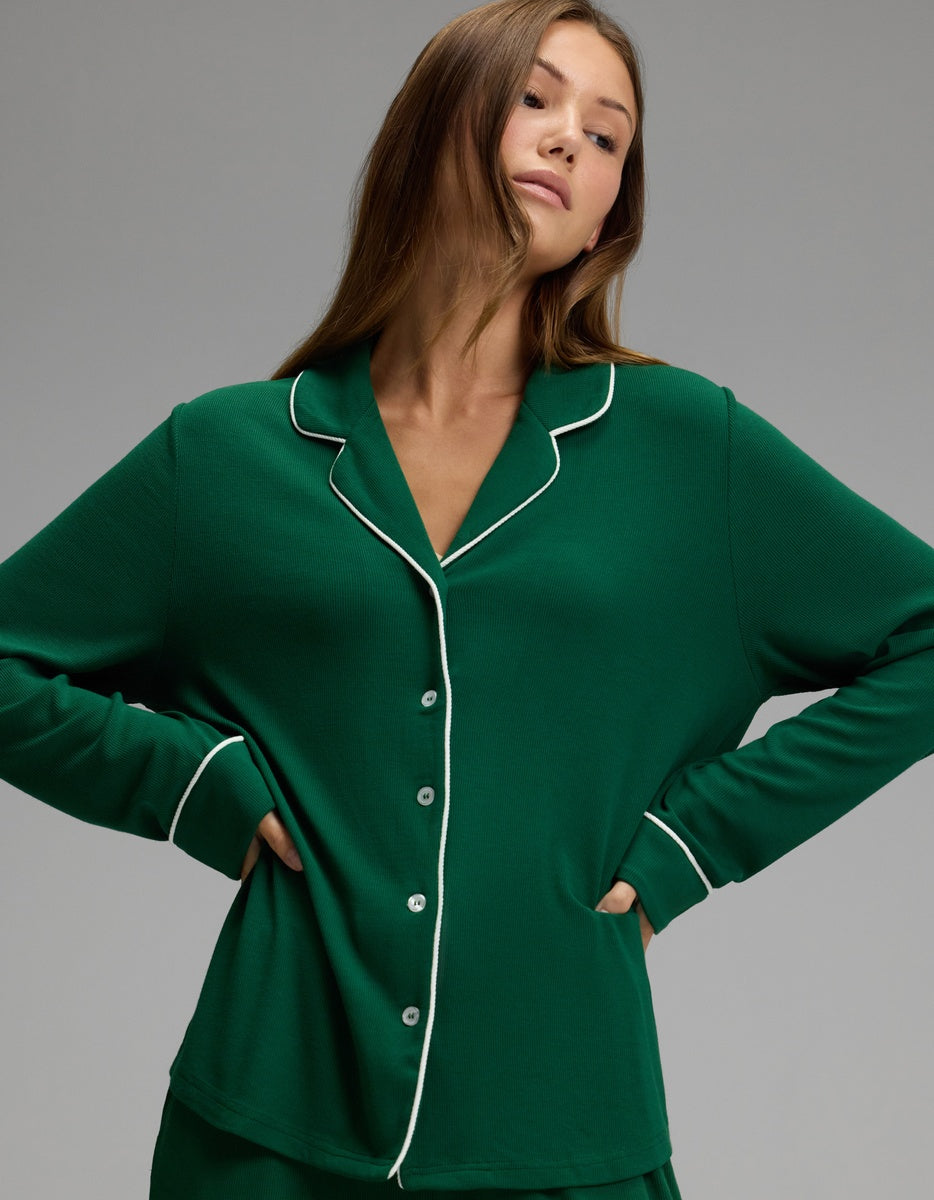 A woman stands with hands on hips, facing slightly to the side against a plain gray background, wearing the Cozy Earth Women’s Bamboo Rib-Knit Classic Long Sleeve Pajama Top in green with white piping.