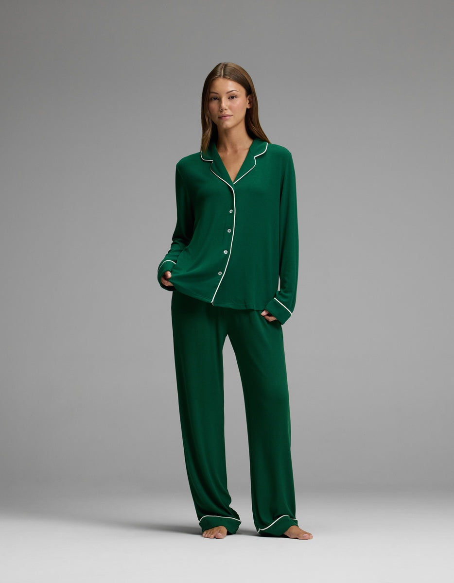 A woman with straight brown hair stands barefoot in a studio, wearing the Cozy Earth Women’s Bamboo Rib-Knit Classic Long Sleeve Pajama Top in dark green with white piping, paired with matching loose pants. She has a neutral expression.
