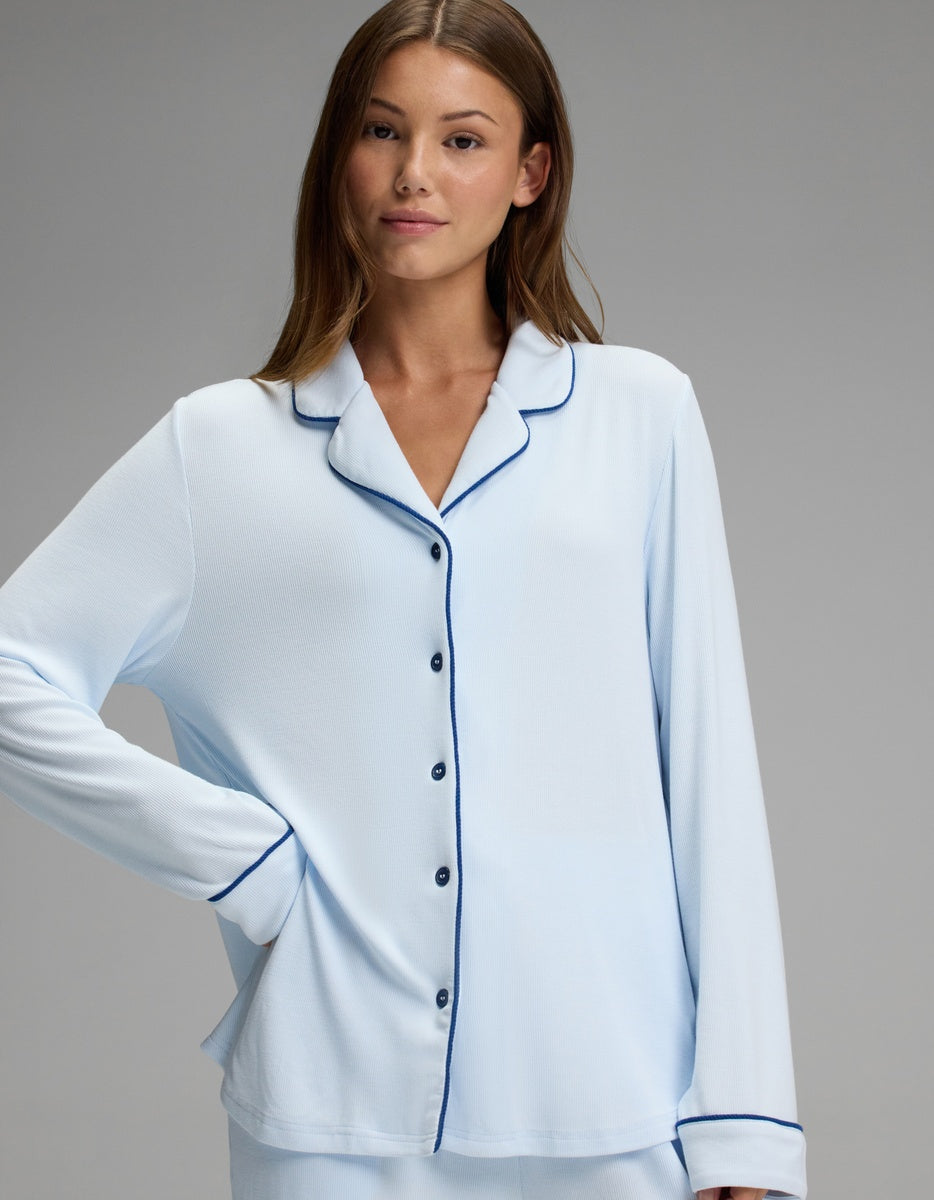 A woman models the Cozy Earth Women’s Bamboo Rib-Knit Classic Long Sleeve Pajama Top in light blue with dark trim, standing with one hand on her hip and looking at the camera against a plain gray background.