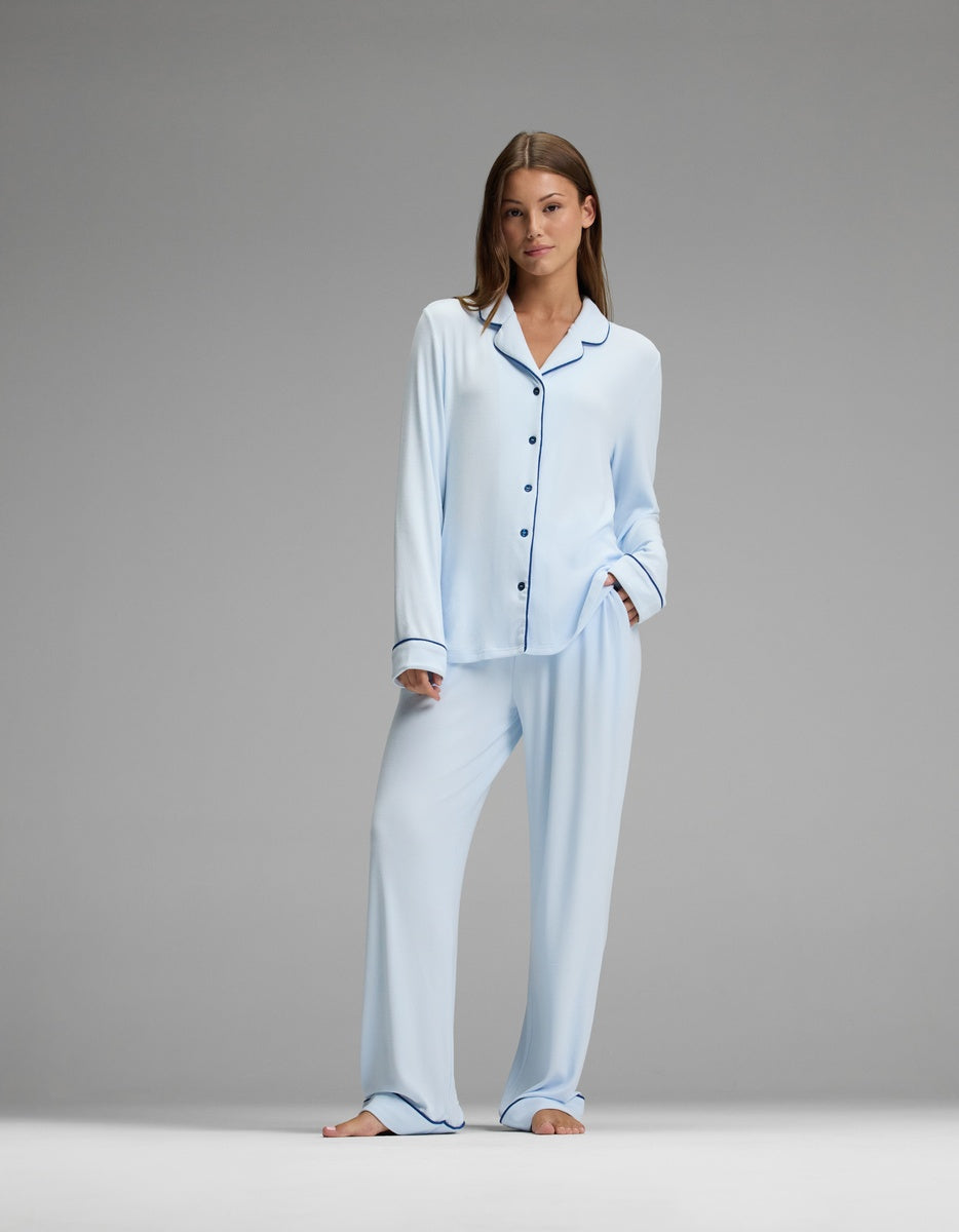 A woman stands barefoot against a plain gray background, looking relaxed with one hand in her pocket. She wears the Cozy Earth Women’s Bamboo Rib-Knit Classic Long Sleeve Pajama Top in light blue with black piping.