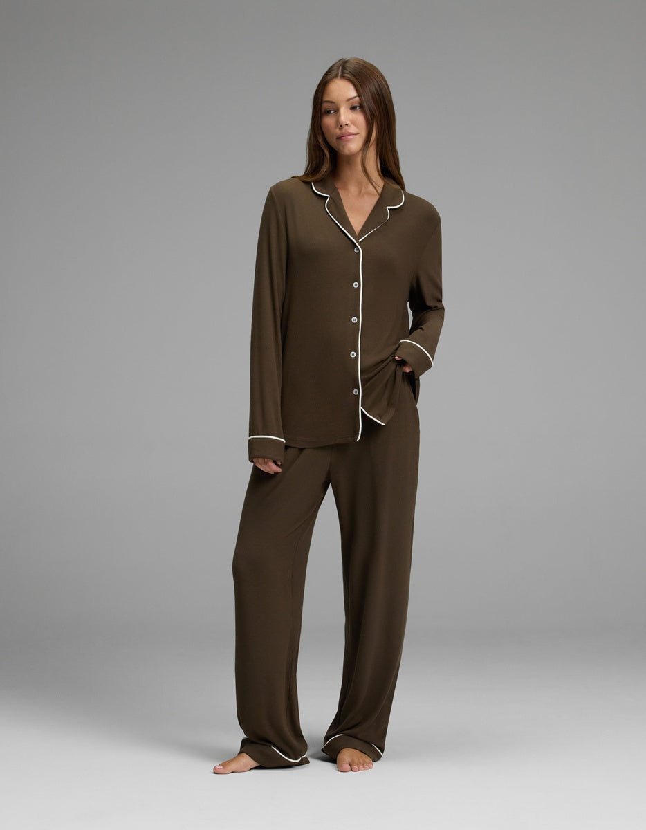 A woman stands barefoot, wearing the Cozy Earth Women’s Bamboo Rib-Knit Classic Long Sleeve Pajama Set—loose brown pajamas with white trim, featuring a button-up top and matching pants—as she poses against a plain gray background.