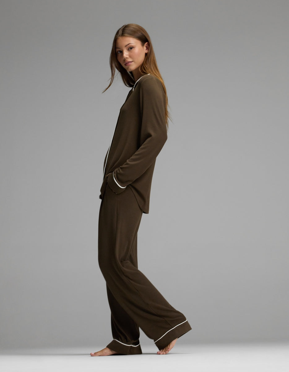 A woman stands barefoot in profile against a plain gray background, wearing the Cozy Earth Women’s Bamboo Rib-Knit Classic Long Sleeve Pajama Set, featuring a matching brown color with white piping on the collar, cuffs, and hems.