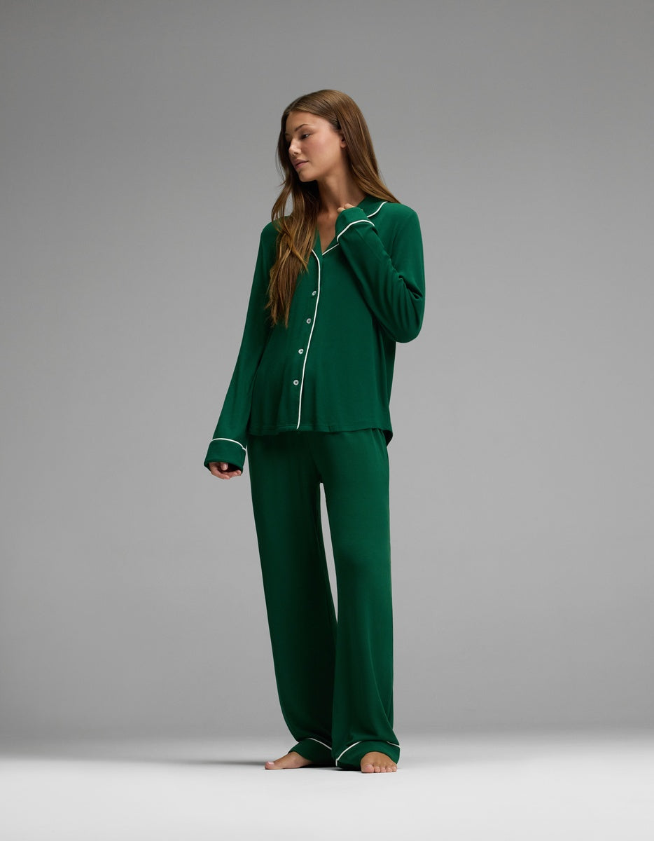A woman stands barefoot, wearing Cozy Earth’s Women’s Bamboo Rib-Knit Classic Long Sleeve Pajama Set in dark green with white trim. The set features a long-sleeve button-up top and wide-leg pants. She gazes to her left against a plain gray background.|Color:Evergreen