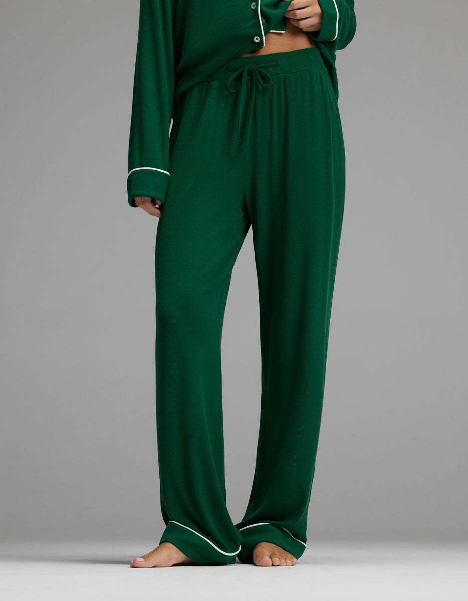 A person is shown standing barefoot on a plain gray background, wearing Cozy Earth’s Women’s Bamboo Rib-Knit Classic Pajama Pant in dark green with a matching top featuring white trim. The individual’s head is not visible in the image.