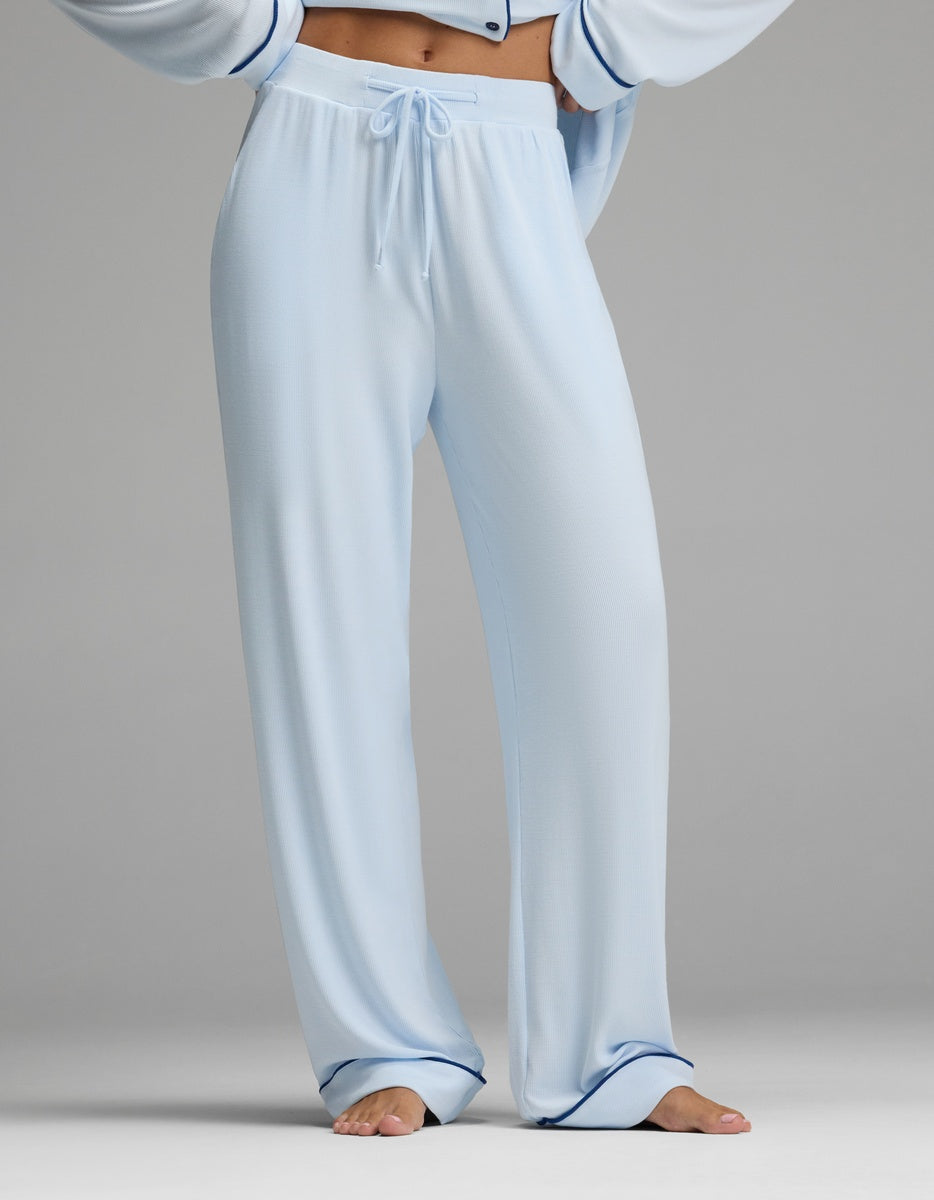 A person is shown wearing the Cozy Earth Women’s Bamboo Rib-Knit Classic Pajama Pant, featuring a loose fit, light blue color, drawstring waist, and dark blue piping at the hems. The upper body and head are not visible, set against a plain gray background.