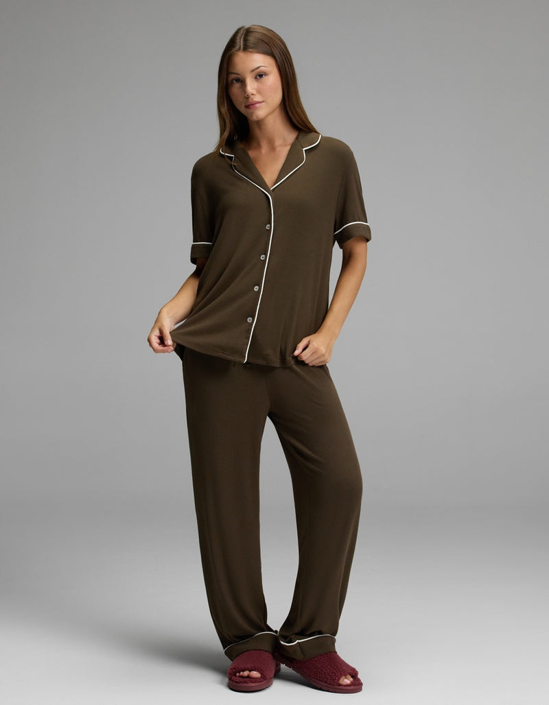 A woman stands against a plain grey background, holding the edge of her Cozy Earth Women’s Bamboo Rib-Knit Classic Short Sleeve Pajama Top in brown with white piping, and wears red slippers.