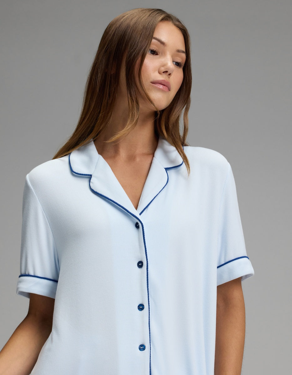 A woman with long brown hair wears the Cozy Earth Women’s Bamboo Rib-Knit Classic Short Sleeve Pajama Top in light blue, featuring dark piping, as she poses against a plain gray background.