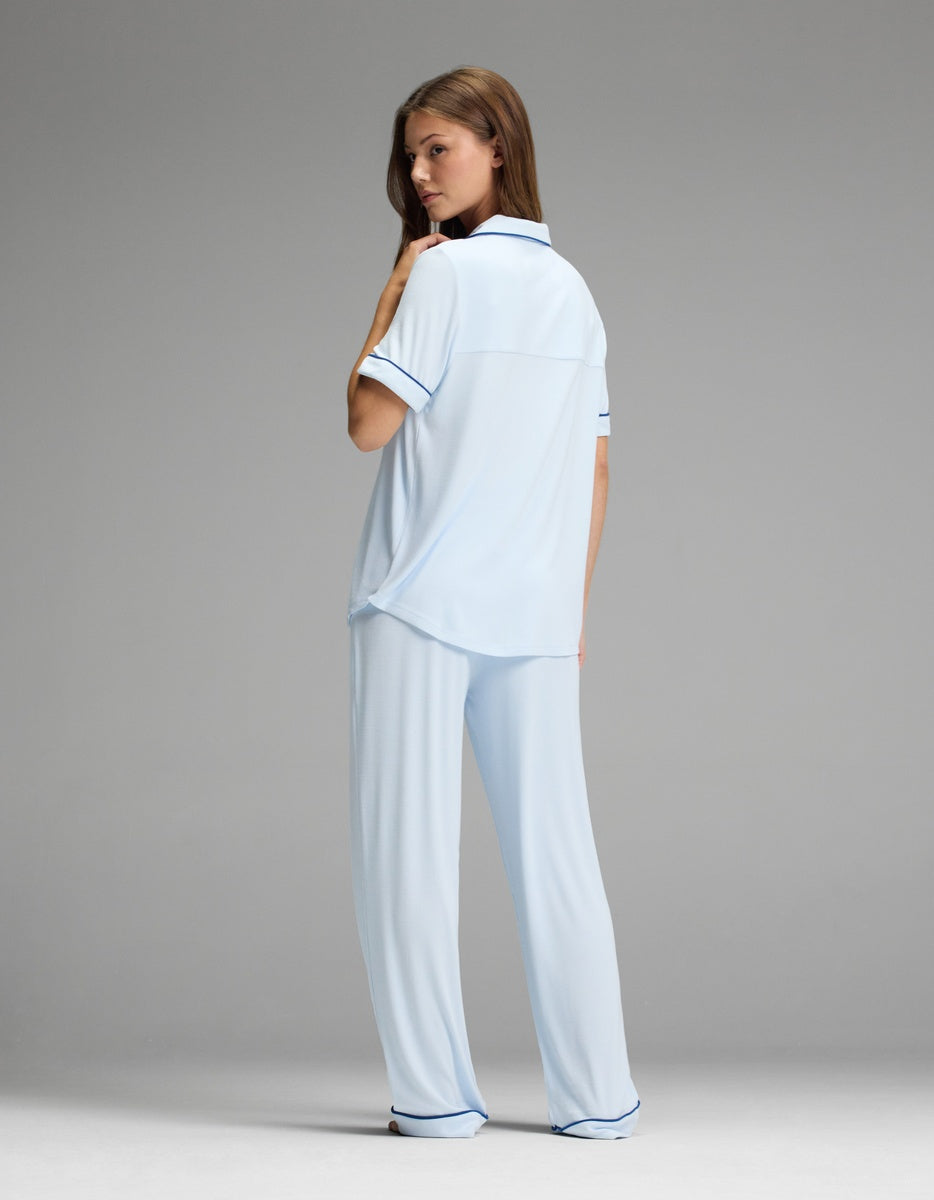 A woman stands against a plain gray background, looking over her shoulder and wearing the Cozy Earth Women’s Bamboo Rib-Knit Classic Short Sleeve & Pant Pajama Set in light blue with dark trim.