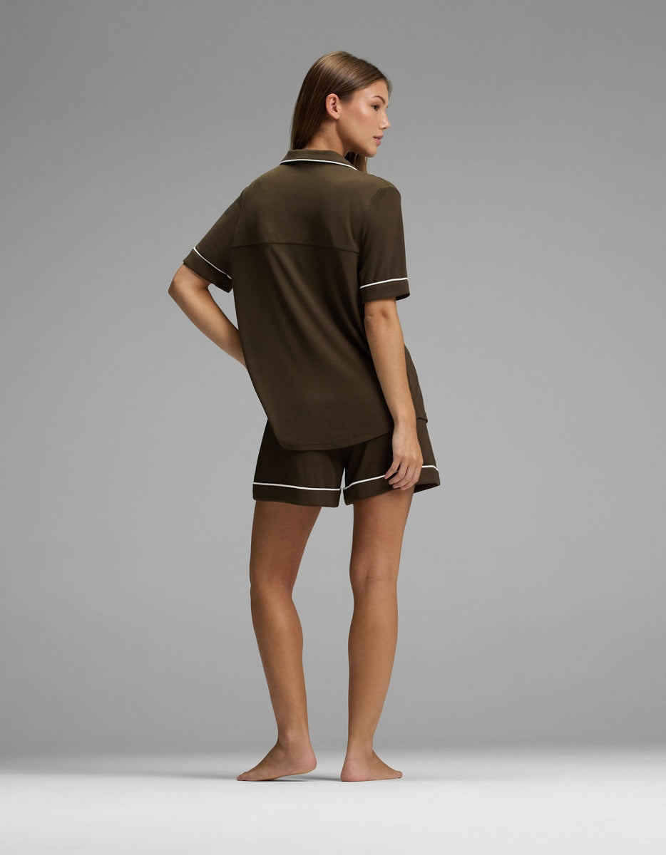 A person stands barefoot on a plain gray surface, facing away, wearing the Cozy Earth Women’s Bamboo Rib-Knit Classic Short Sleeve Pajama Set in dark brown with light trim.|Color:Espresso