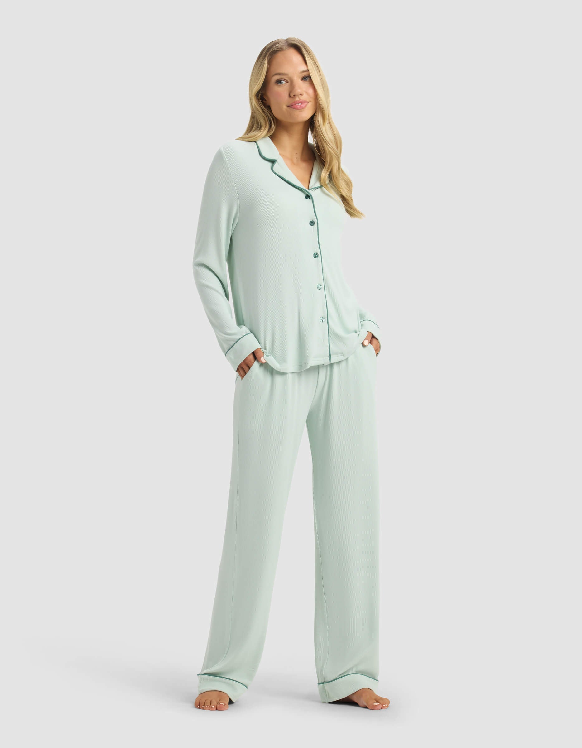 A woman poses barefoot before a plain light background, smiling softly in Cozy Earth’s Women’s Bamboo Rib-Knit Classic Long Sleeve Pajama Set—light blue with button-up top, wide-leg pants, and her hands in the pockets.|Color:Seafoam