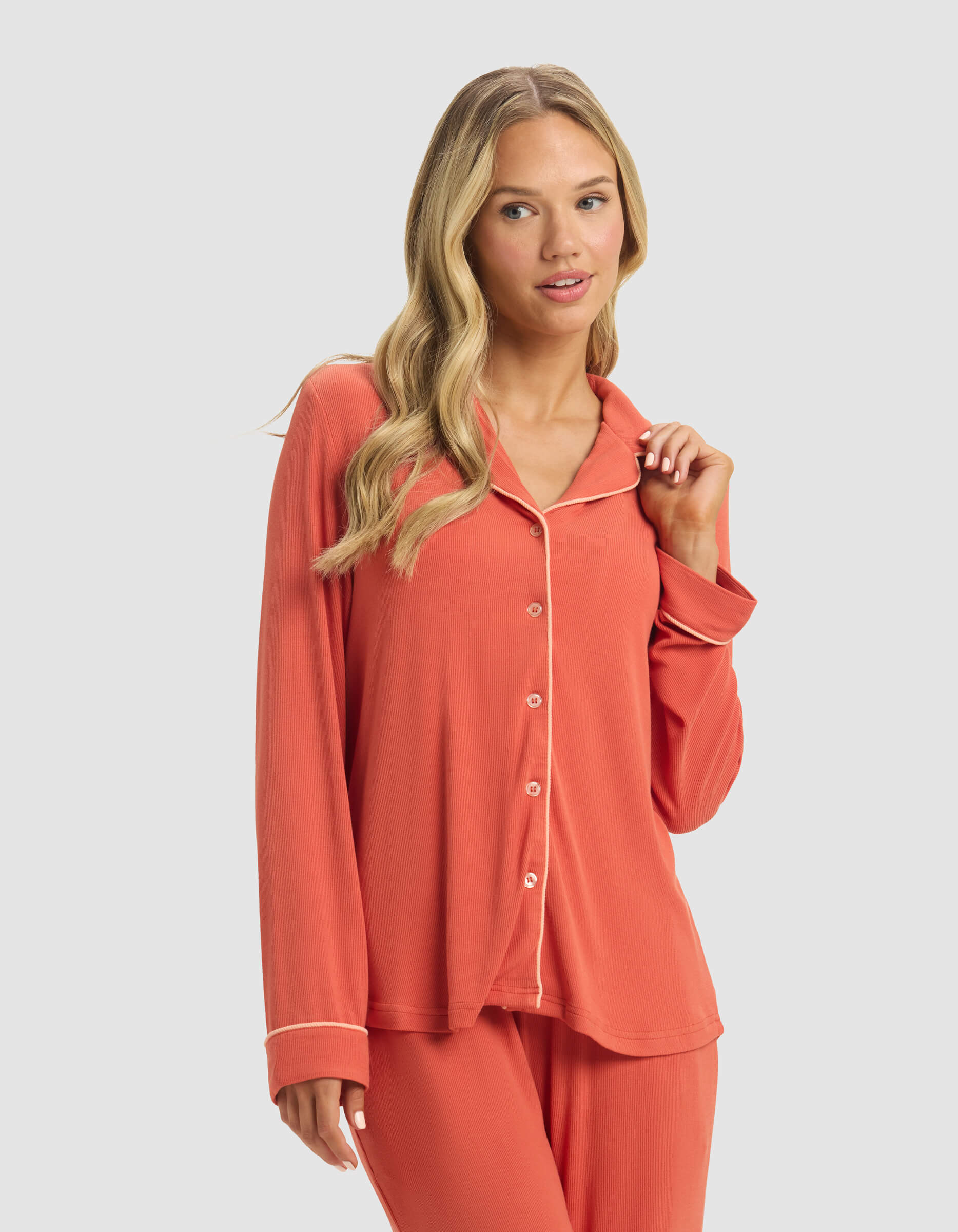 A woman with long blonde hair wears the Cozy Earth Women's Bamboo Rib-Knit Classic Long Sleeve Pajama Top in coral with white piping, paired with matching pants, standing against a plain light gray background.|Color:Sunset