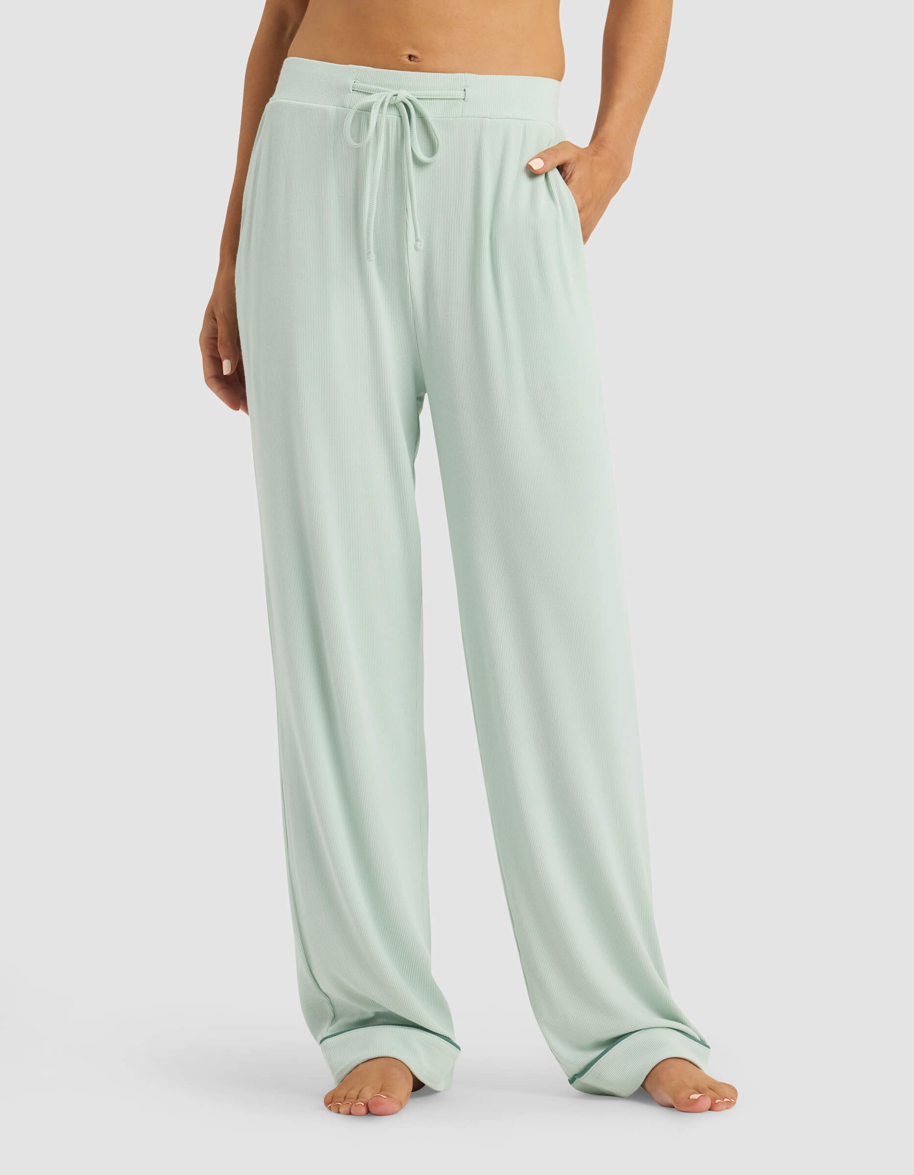 A person stands barefoot with hands in the pockets, wearing Cozy Earth Women's Bamboo Rib-Knit Classic Pajama Pant in light green. Their upper body is cropped out of the image.
