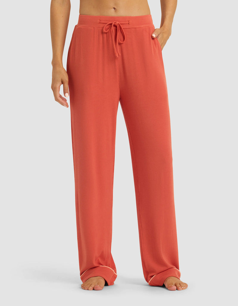A person stands barefoot against a plain light background wearing Cozy Earth's Women's Bamboo Rib-Knit Classic Pajama Pant in a loose, coral-colored style with a drawstring waist.