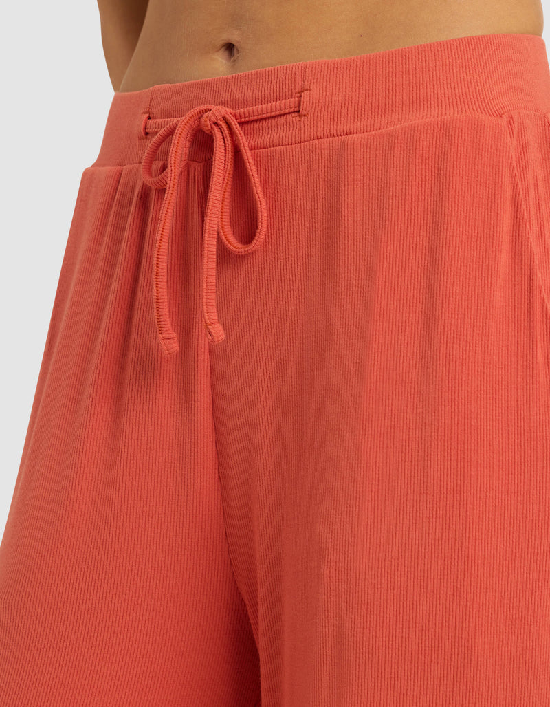 Close-up of a person wearing Cozy Earth Women's Bamboo Rib-Knit Classic Pajama Pant in coral, featuring a high waist, ribbed texture, and drawstring tie. Only the lower torso and upper legs are shown; fabric looks soft and comfortable.