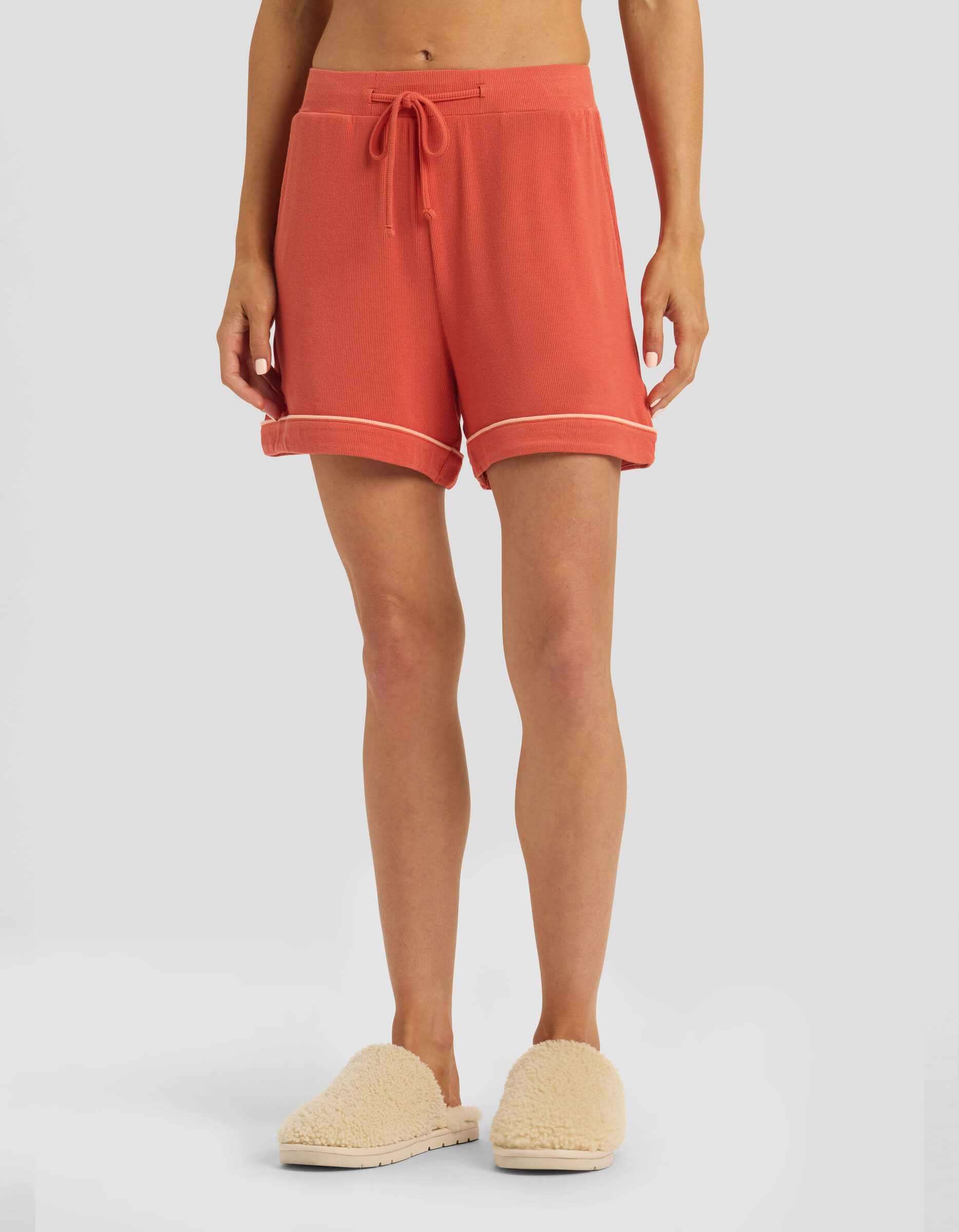 A person is seen from the waist down wearing Cozy Earth Women's Bamboo Rib-Knit Classic Pajama Shorts in coral, featuring a drawstring and rolled hems, paired with cream fuzzy slippers against a plain light gray background.|Color:Sunset