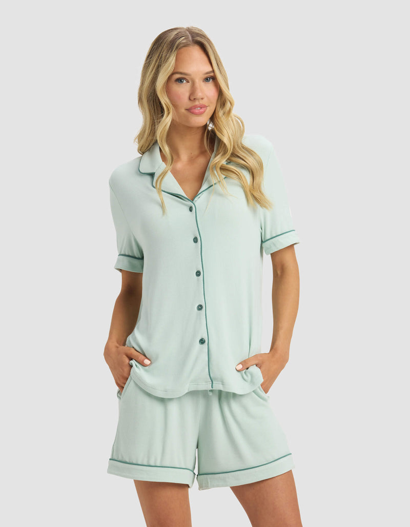 A woman with long, wavy blonde hair wears the Cozy Earth Women's Bamboo Rib-Knit Classic Short Sleeve Pajama Top in light green, paired with matching shorts. She stands against a plain light background with her hands in her pockets.