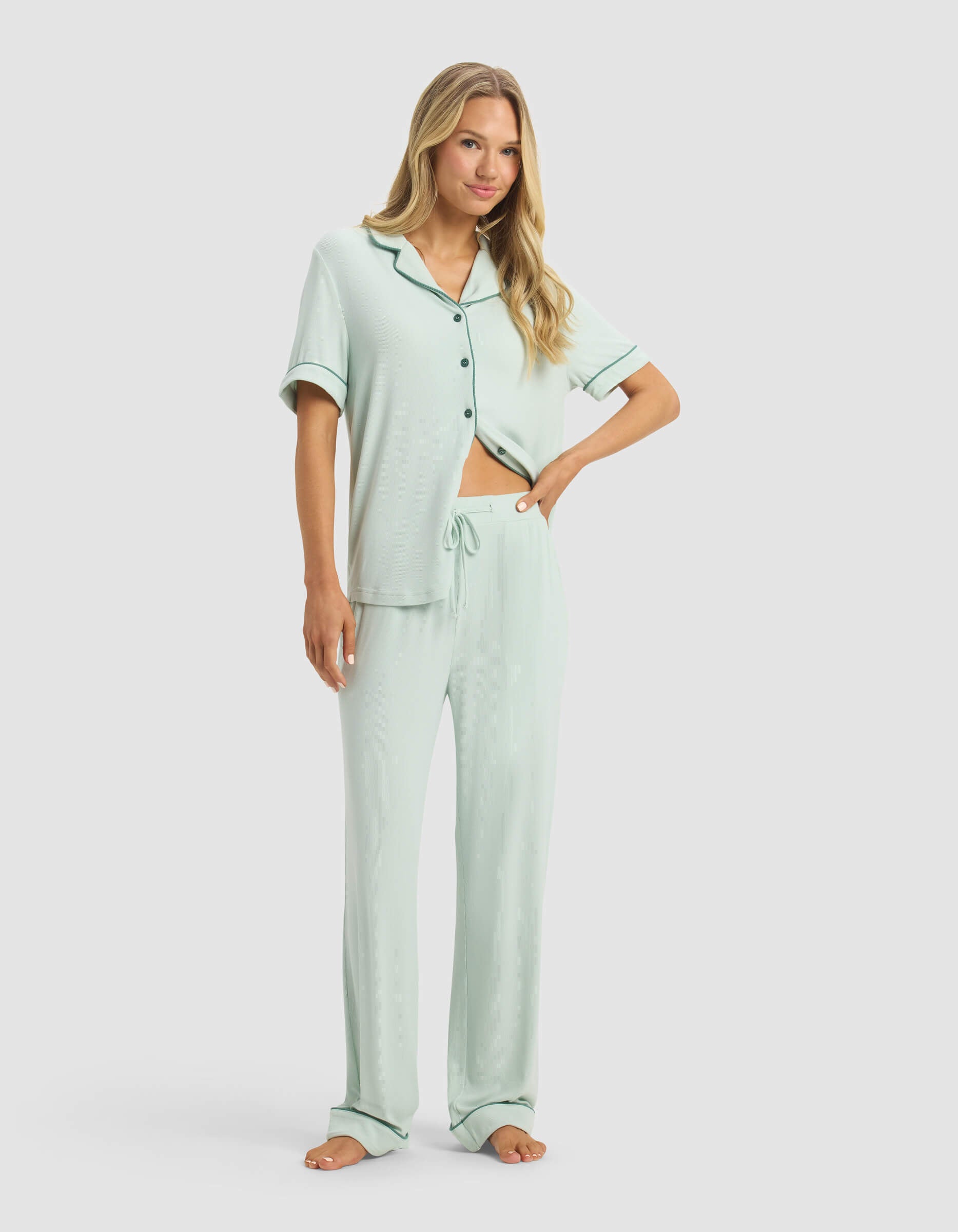 A woman with long blonde hair stands barefoot, smiling slightly, in front of a plain light background wearing Cozy Earth's Women's Rib-Knit Classic Short Sleeve & Pant Pajama Set—a light green, short-sleeve button-up top with loose-fitting pants.