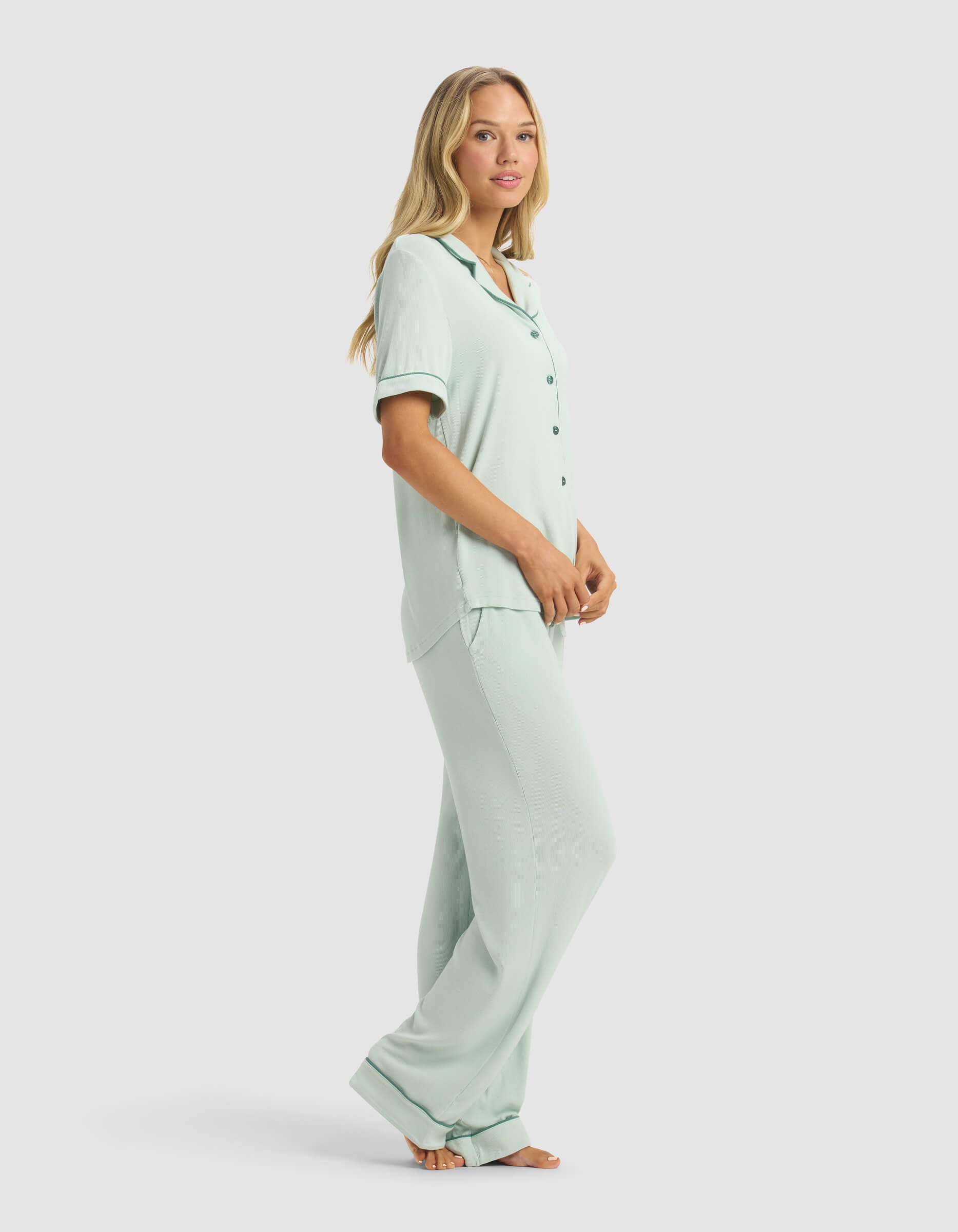 A woman stands barefoot in the Cozy Earth Women's Rib-Knit Classic Short Sleeve & Pant Pajama Set, featuring a light blue button-up top and pants. She poses side-on, looking at the camera against a plain light background.|Color:Seafoam