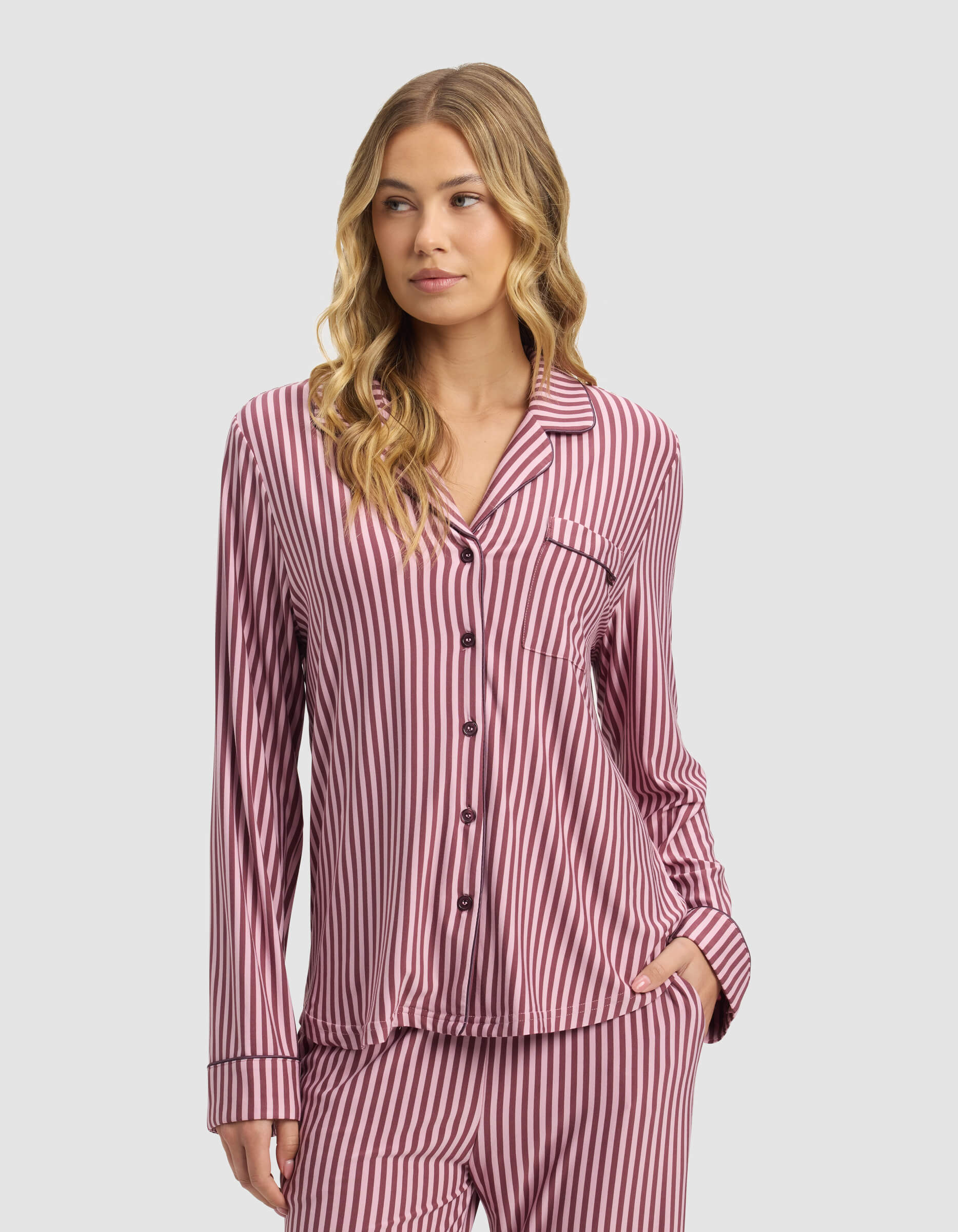 A woman with long wavy blonde hair wears the Cozy Earth Women's Bamboo Stretch-Knit Long Sleeve Pajama Top in pink and white stripes, paired with matching pants, standing against a plain light gray background.|Color:Market Stripe in Mulberry
