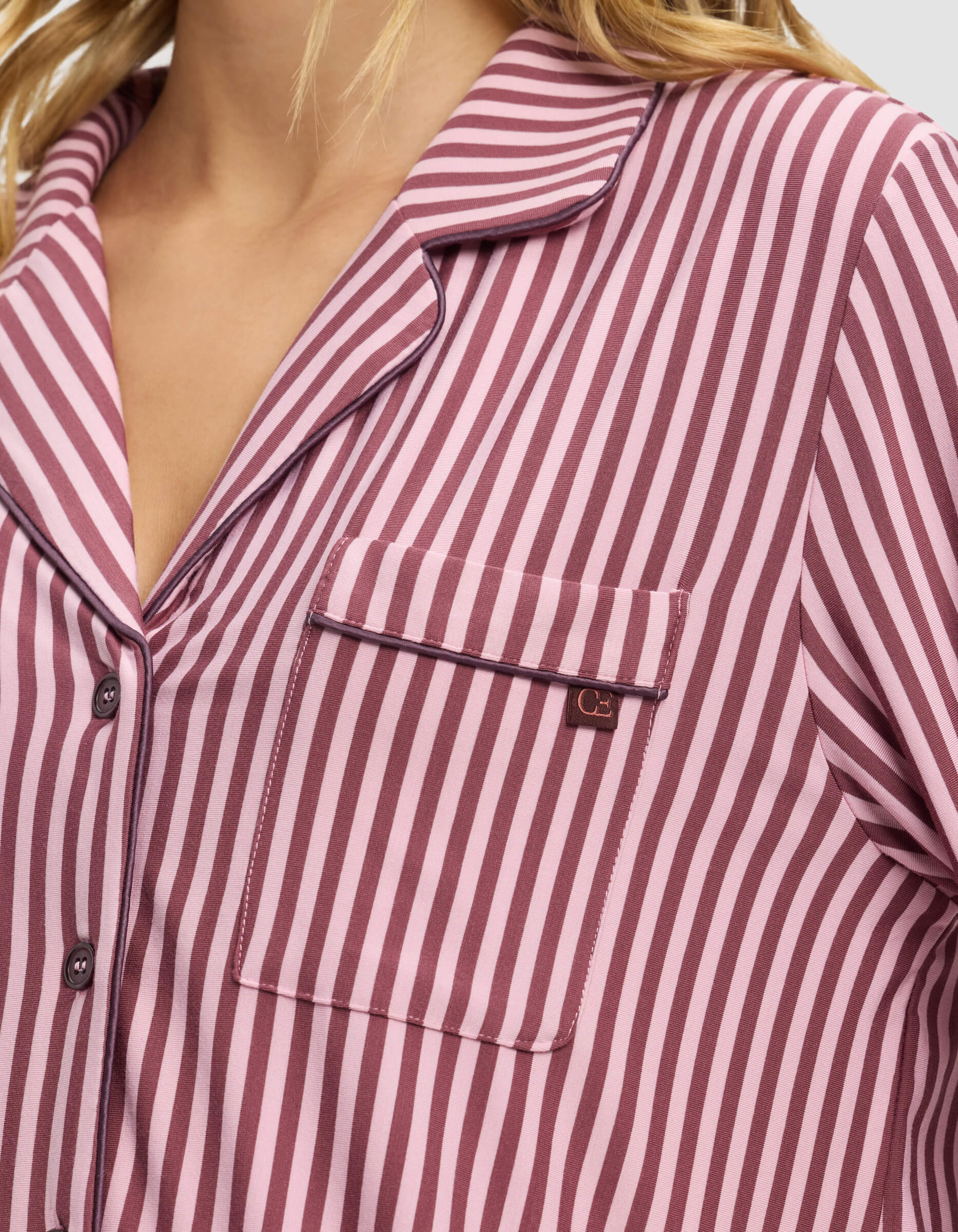 A close-up of a person wearing the Cozy Earth Women's Bamboo Stretch-Knit Long Sleeve Pajama Top in pink and burgundy stripes, featuring a front chest pocket with a small metal tag and a collared, button-up design.|Color:Market Stripe in Mulberry
