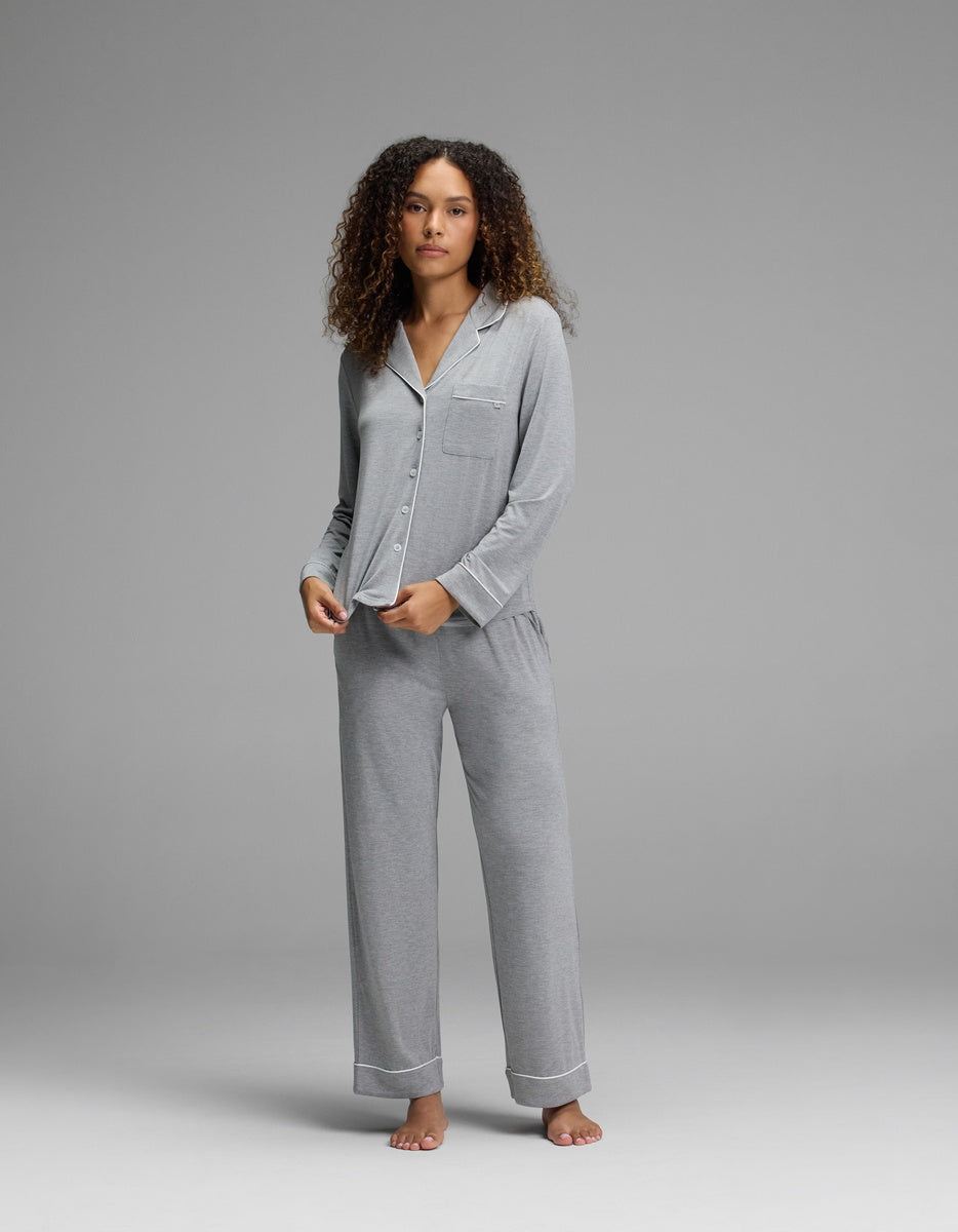 A person with curly hair stands barefoot on a gray background, wearing the Cozy Earth Women’s Bamboo Stretch-Knit Long Sleeve Pajama Top - Petite in light gray, paired with matching long pants.|Color:Light Heather Grey