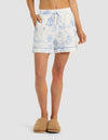A woman wears Cozy Earth’s Women's Bamboo Stretch-Knit Pajama Short in a white and blue floral print with a drawstring waist and rolled hems, paired with tan fuzzy slippers, standing against a plain light background.|Color:Garden Toile in Sea Salt
