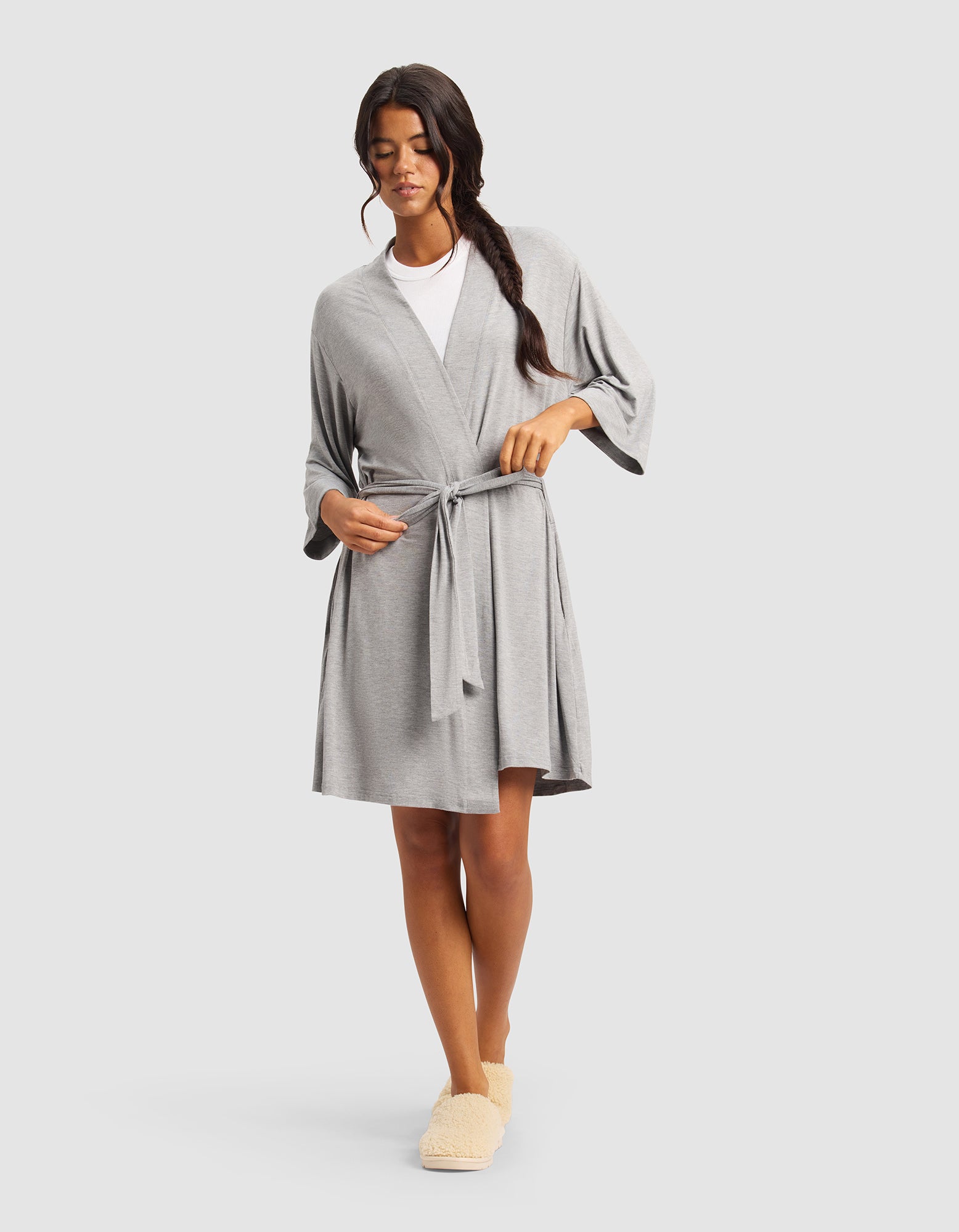 A woman with long dark hair in a side braid wears the Cozy Earth Women's Bamboo Stretch-Knit Kimono Robe, tied at the waist over a white top and beige slippers, standing against a plain light gray background.