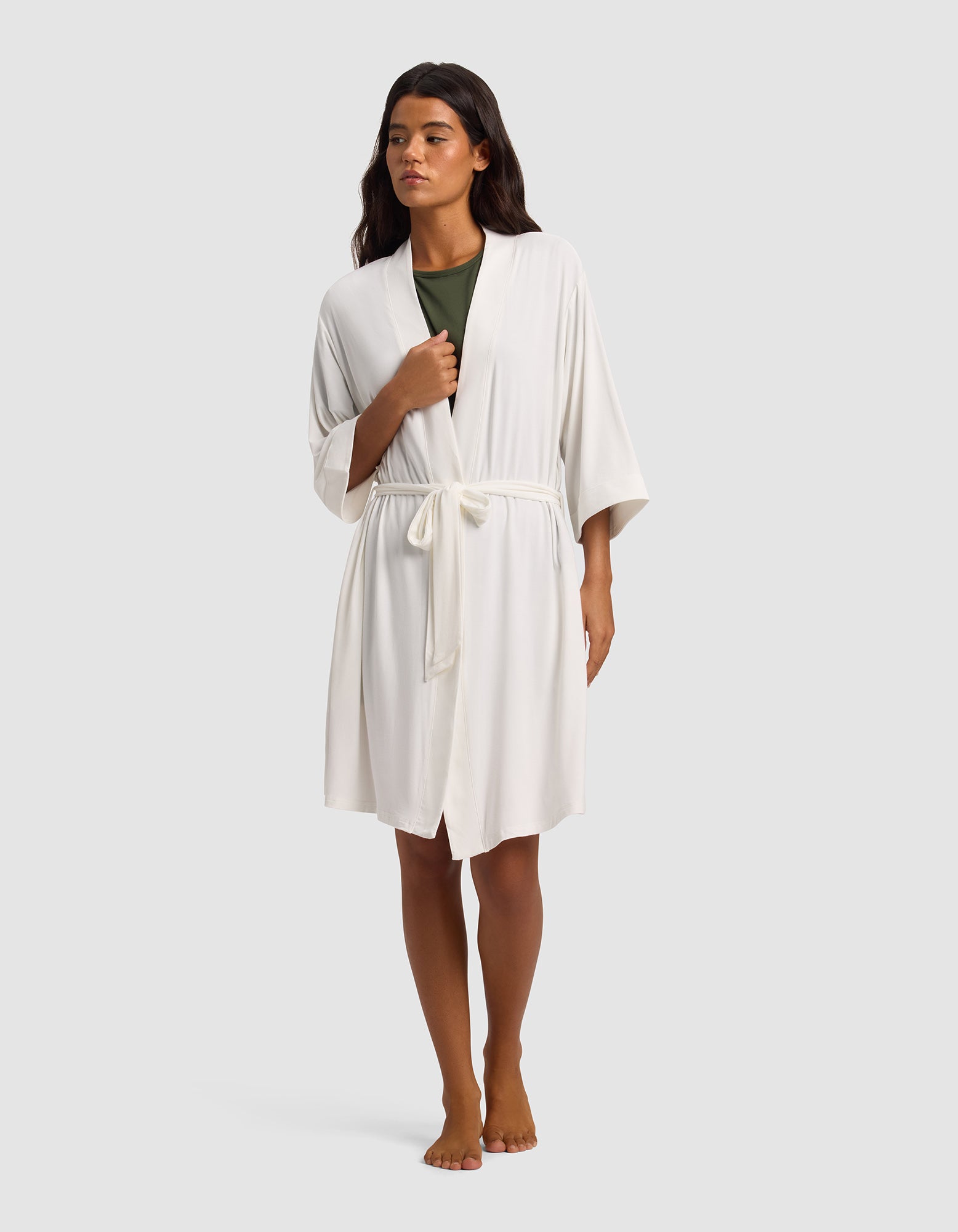 A person stands barefoot against a plain light gray background, wearing Cozy Earth's Women's Bamboo Stretch-Knit Kimono Robe in white with a tie belt over a green top.