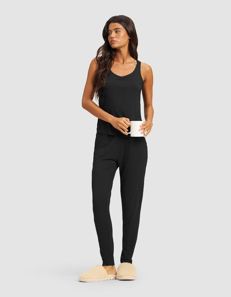 A woman in the Cozy Earth Women's Bamboo Stretch-Knit Tank & Tapered Pant Set, with long brown hair and beige slippers, stands holding a white mug and looking to the side against a plain light gray background.