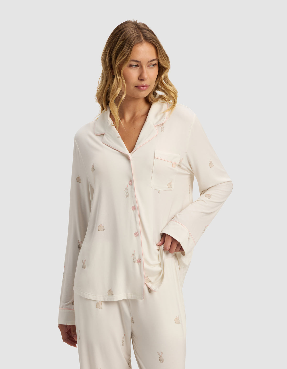 A woman wears the Cozy Earth Women's Bamboo Stretch-Knit Long Sleeve Pajama Top in light cream with a small bunny print and pink piping, standing with one hand in her pocket, smiling slightly against a plain light gray background.