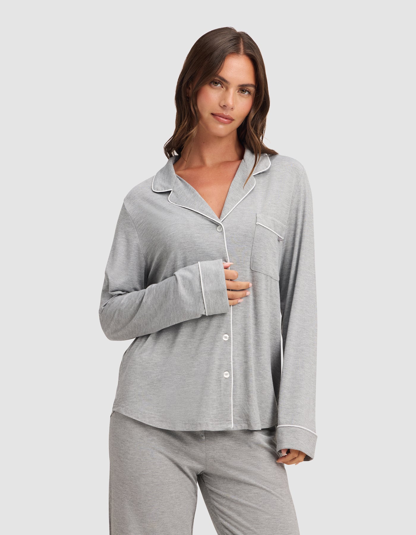 A woman in the HIDE Women's Bamboo Stretch-Knit Long Sleeve Pajama Top, light gray with white piping and buttons, stands against a plain background, softly smiling as she gently holds the top near her chest. 