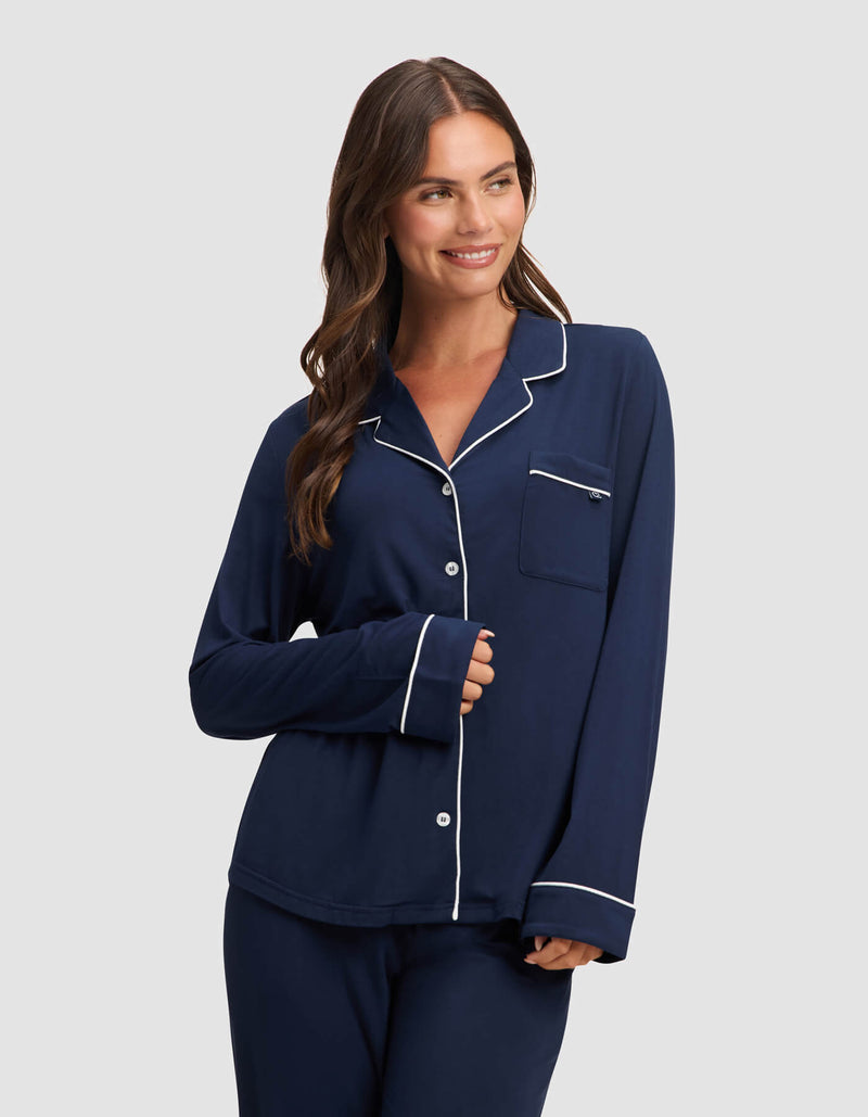 A woman with long brown hair smiles in a Cozy Earth Women's Bamboo Stretch-Knit Long Sleeve Pajama Top, navy blue with white piping, paired with matching pants, against a plain light gray background.