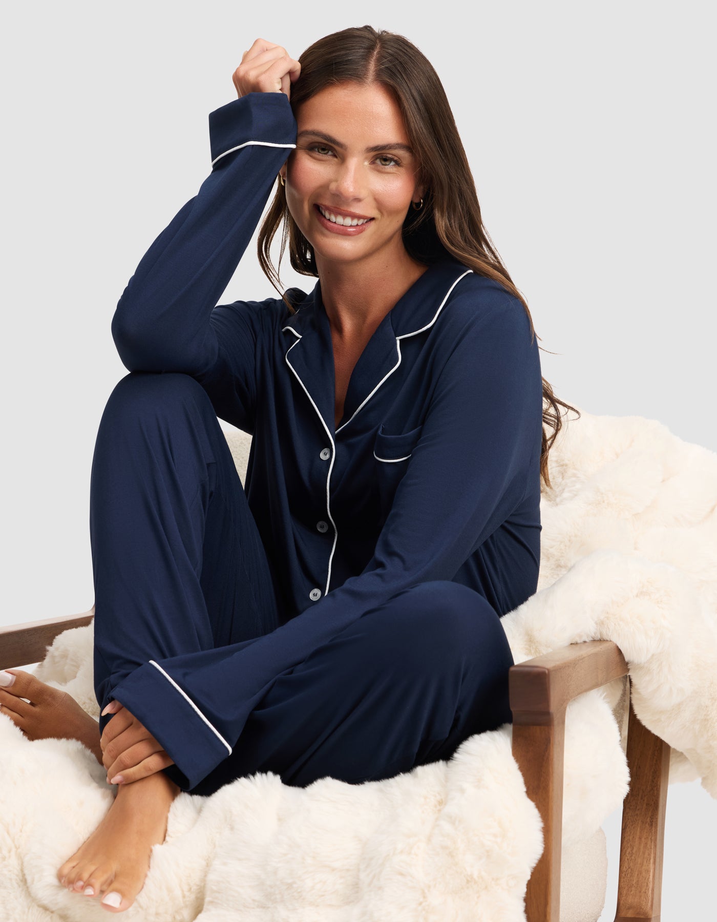 A woman with long brown hair smiles barefoot on a white faux fur chair, wearing the HIDE Women's Bamboo Stretch-Knit Long Sleeve Pajama Top in navy blue with white piping, set against a light gray background. 