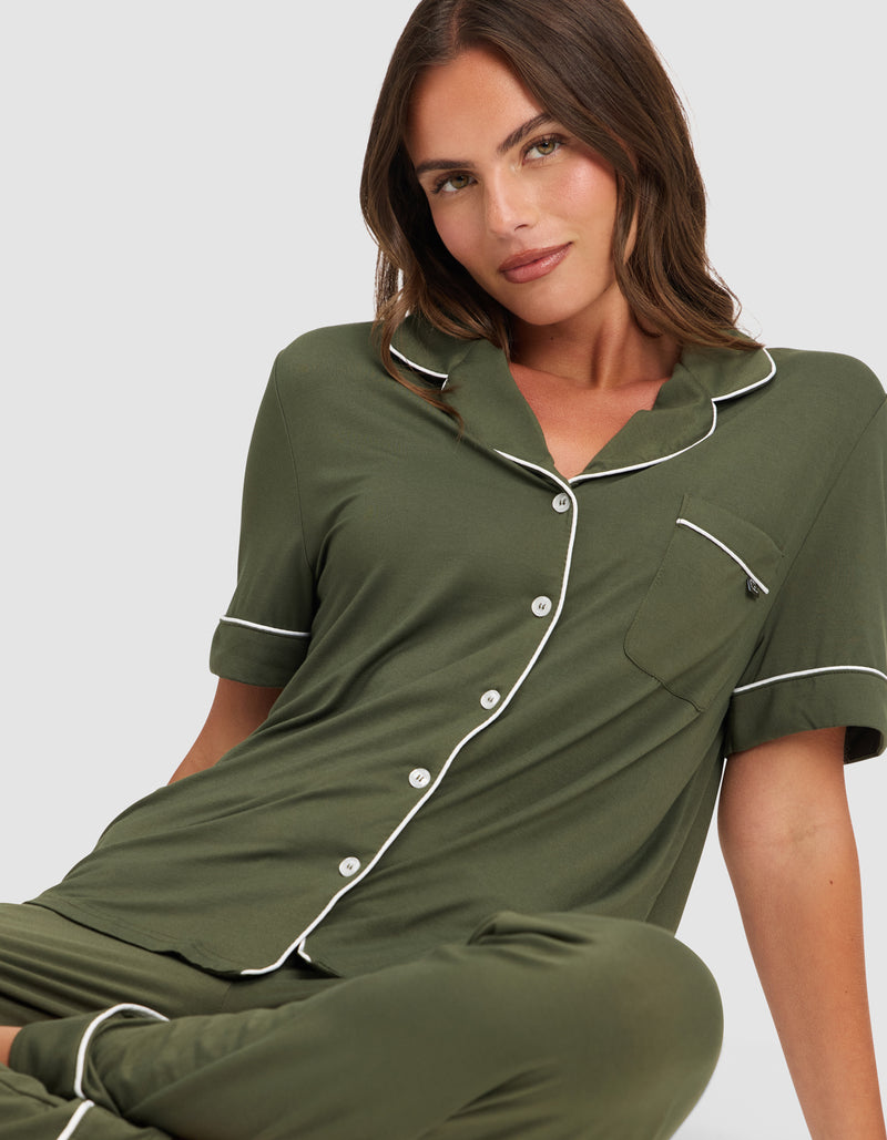 A woman with brown hair wears the HIDE Women's Bamboo Stretch-Knit Short Sleeve Pajama Top in olive green with white piping and buttons. She sits on the floor, facing the camera with a slight smile against a plain light background.