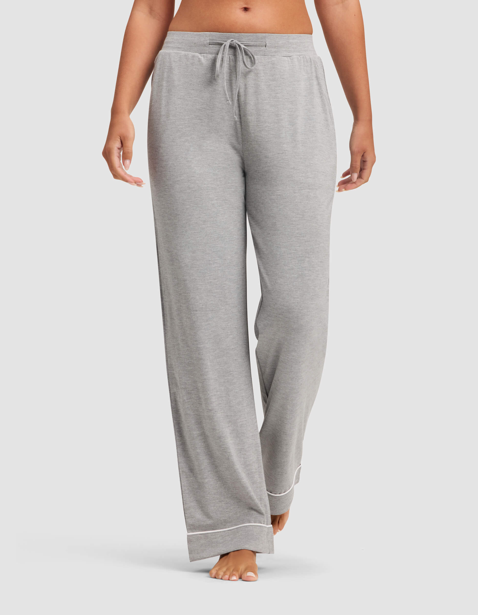 A woman wears Cozy Earth Women's Bamboo Stretch-Knit Pajama Pants in light gray, featuring a drawstring waist and white trim at the cuffs, standing barefoot against a plain background.