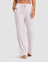 A woman models the Cozy Earth Women's Bamboo Stretch-Knit Pajama Pant in light pink, featuring a drawstring waist and pockets, while standing barefoot against a plain light background.|Color:Lilac