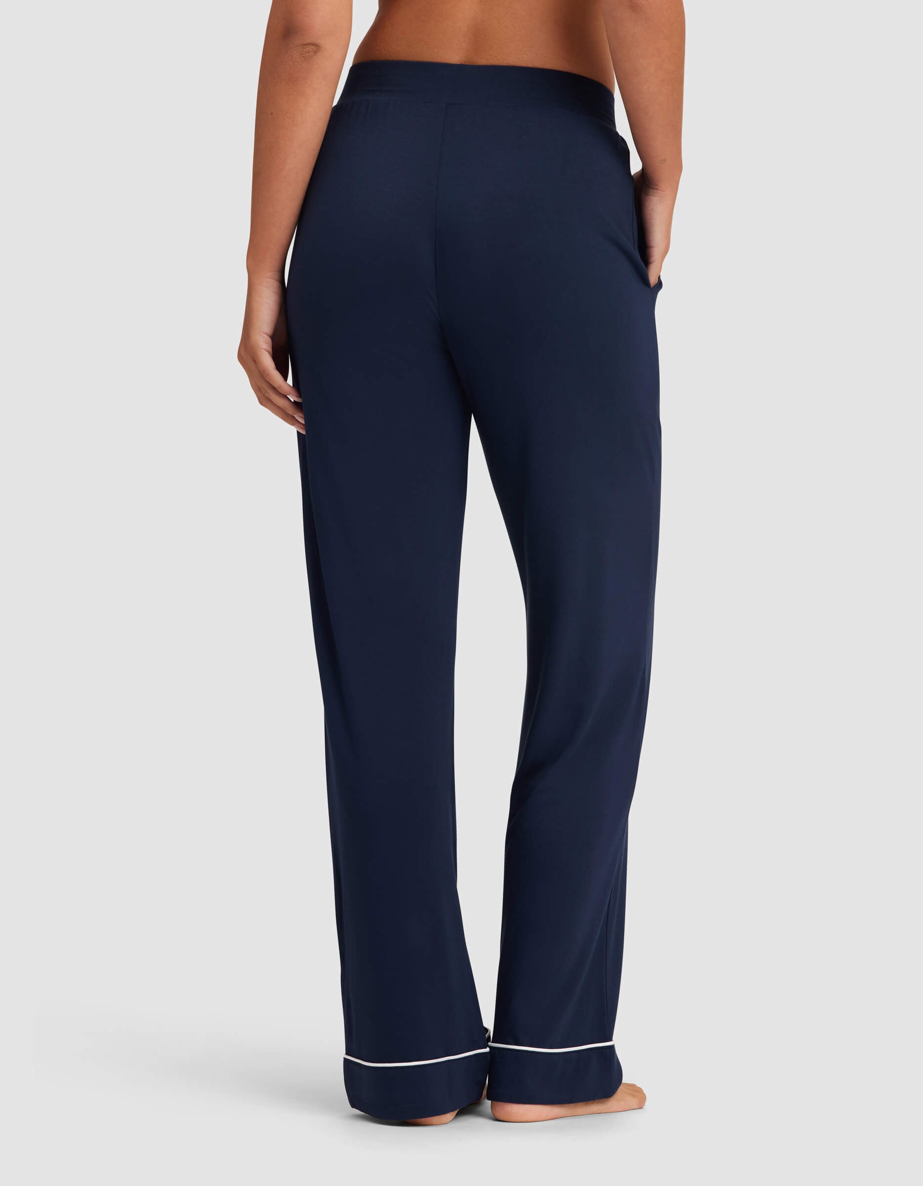 A woman stands barefoot facing away, wearing Cozy Earth Women's Bamboo Stretch-Knit Pajama Pant in navy with white piping at the cuffs and one hand in a pocket against a plain light gray background.|Color:Navy